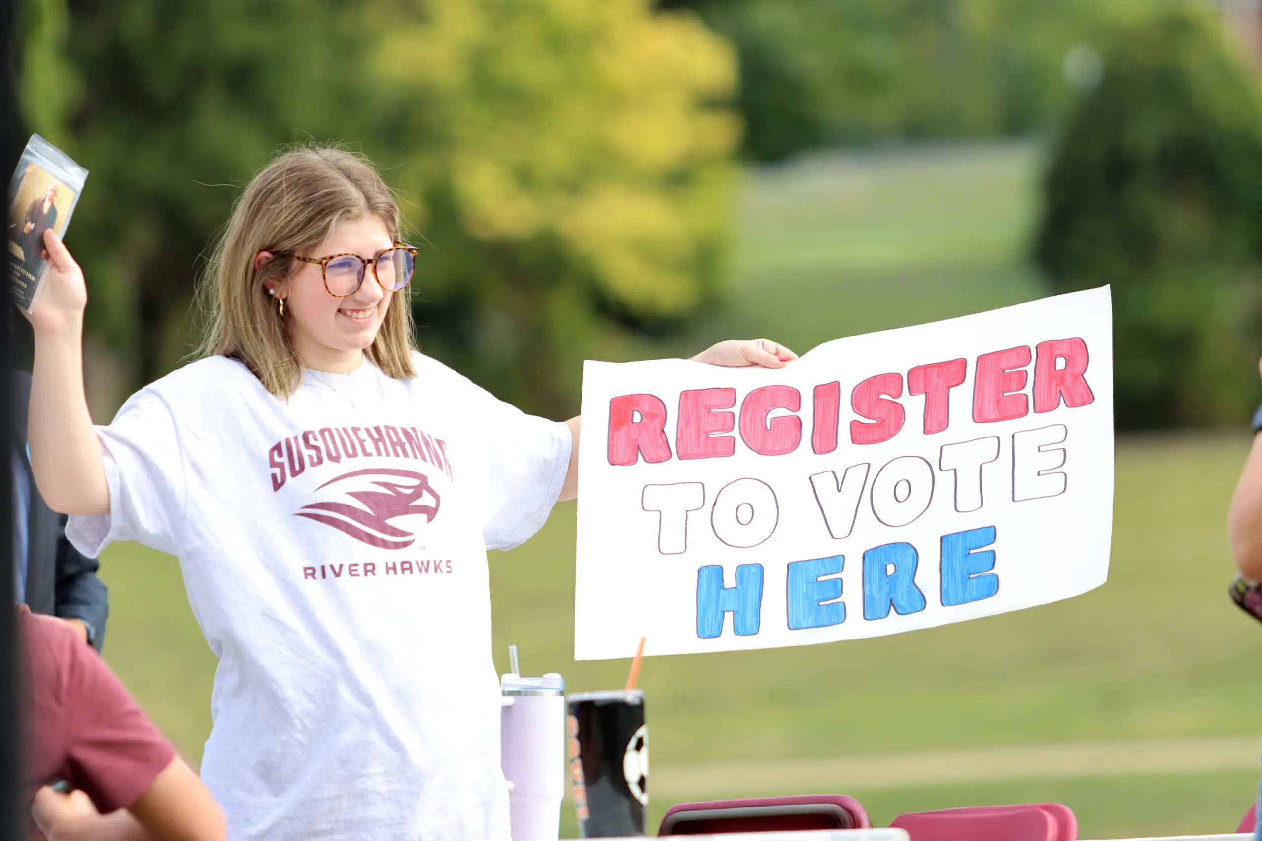 A person wearing glasses and a "Susquehanna River Hawks" shirt holds a sign that reads "Register to Vote Here" in red and blue letters. They are outside, with trees and grass in the background.