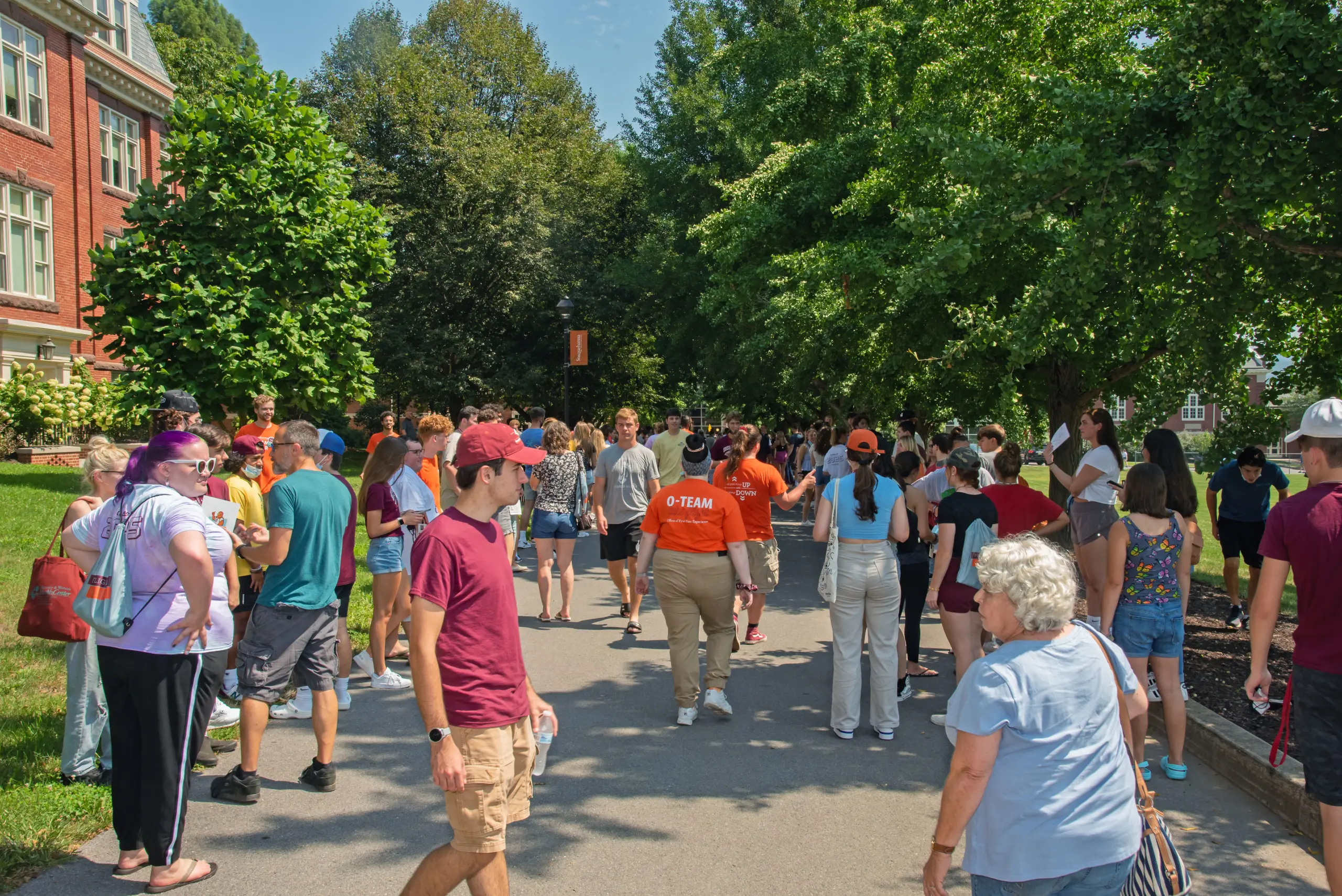A diverse crowd of people walks down a tree-lined path on a sunny day. Some individuals wear orange shirts with "O-Team" printed on the back. A brick building and green trees are visible in the background.