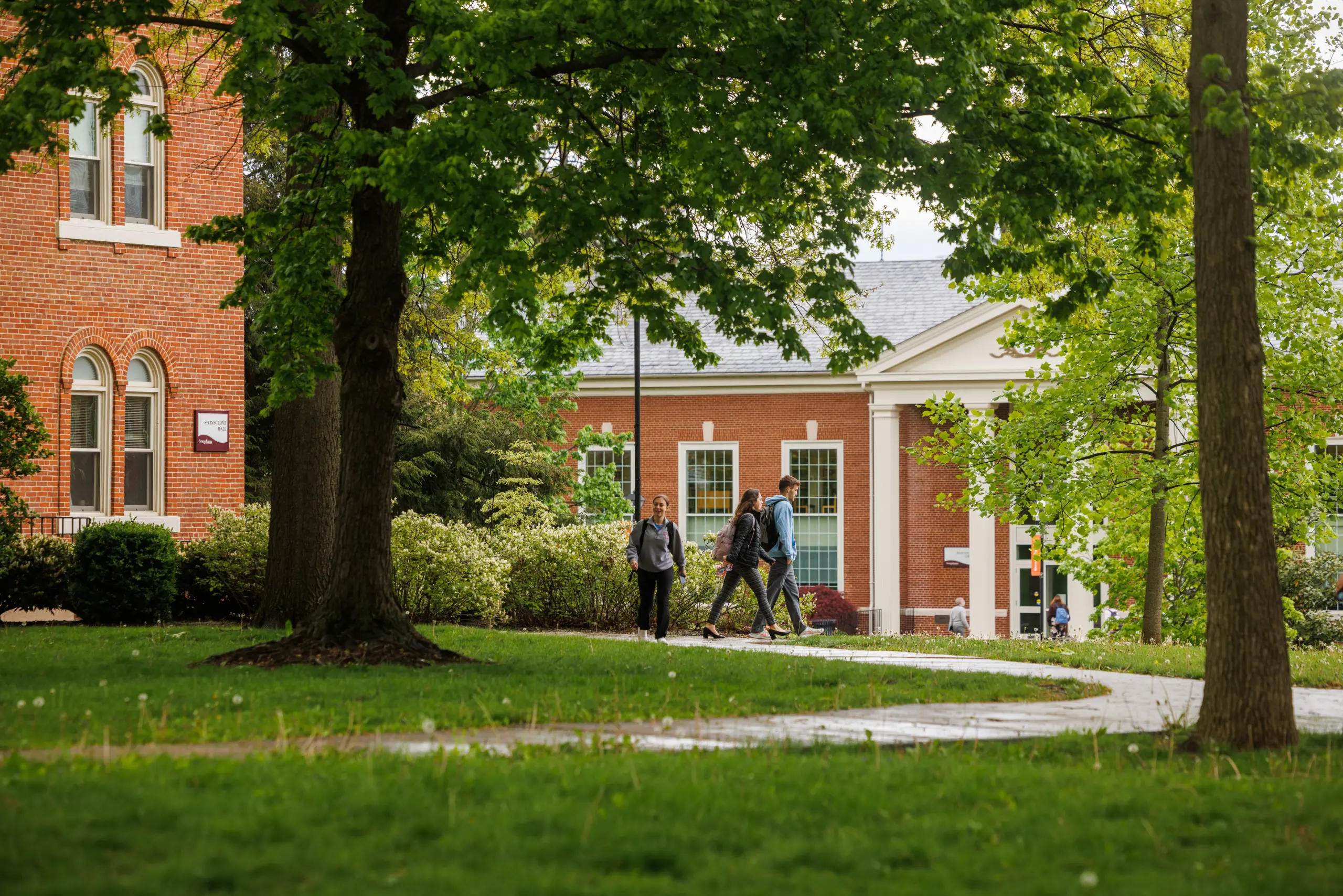 A group of people walk along a path in a college campus setting, surrounded by lush green trees and grass. Brick buildings with large windows are visible in the background, evoking a peaceful and academic atmosphere.