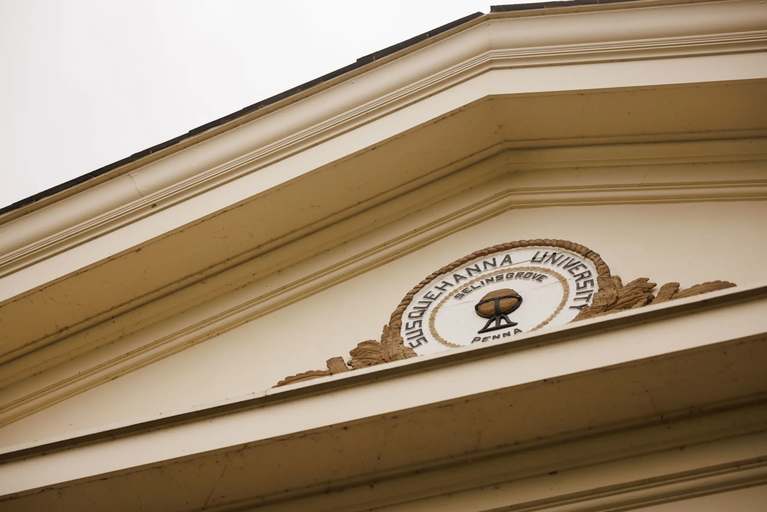 A triangular architectural pediment with the seal of Susquehanna University. The seal features a globe and text reading "Susquehanna University" and "Penna." The design is set against a cloudy sky backdrop.