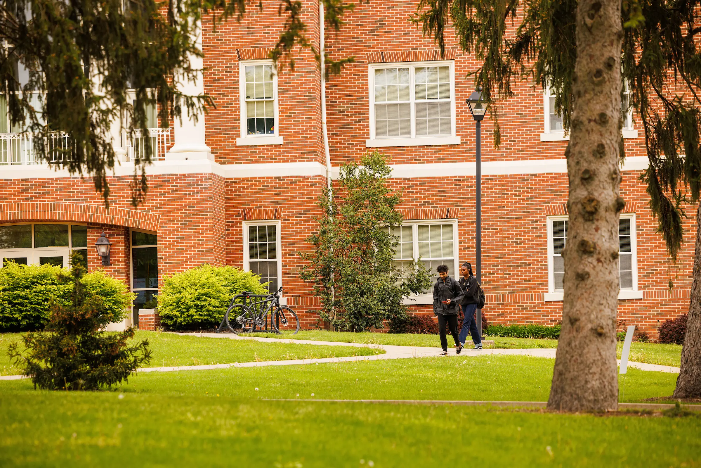 A red brick building with white-framed windows stands against a green lawn and shrubbery. Two people walk on a path beside a bike rack. A tree in the foreground partially obscures the view.