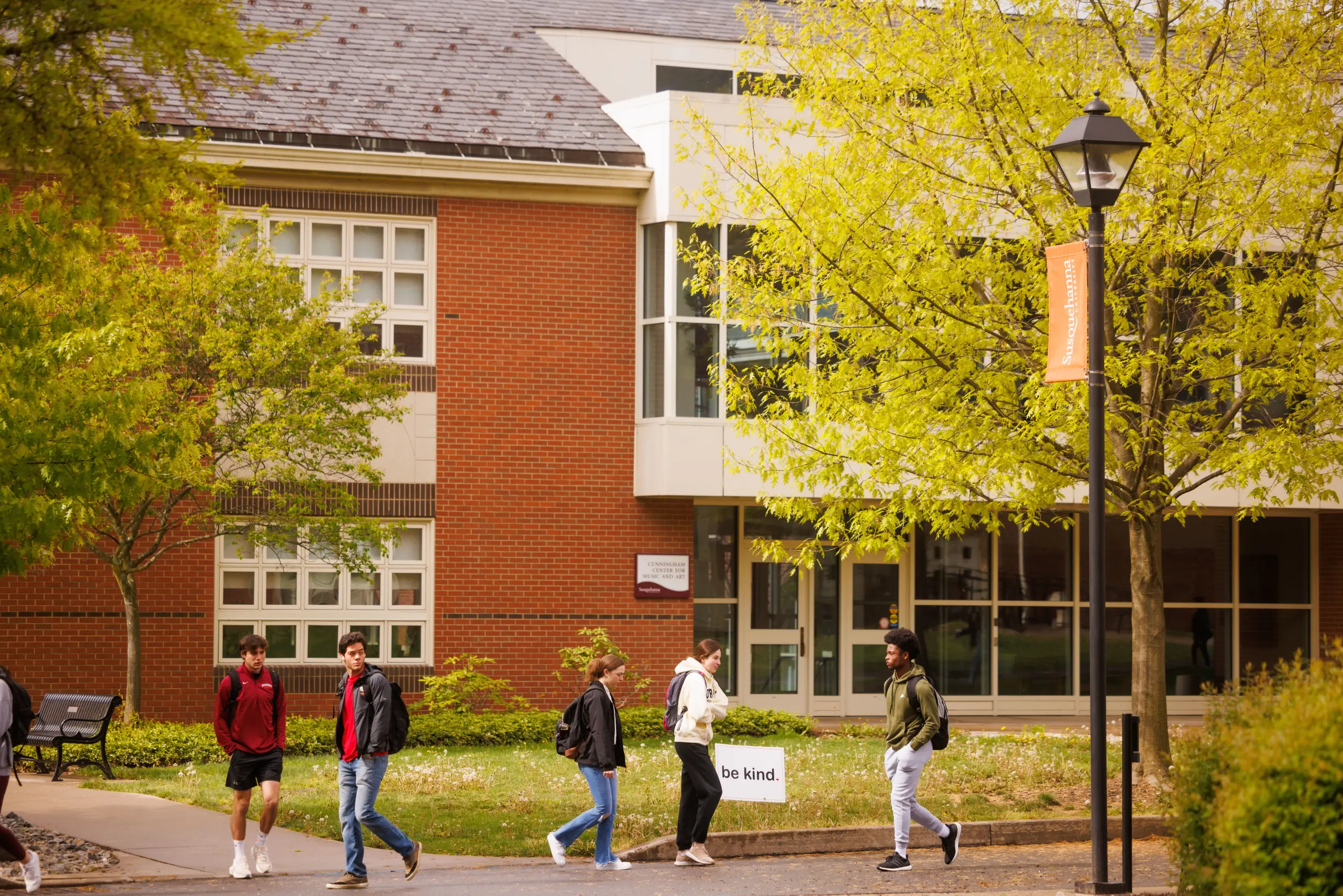 Students walking on a college campus path, surrounded by green trees. A brick building with many windows is in the background. One student holds a white sign with the words "be kind." A lamp post with an orange banner is also visible.