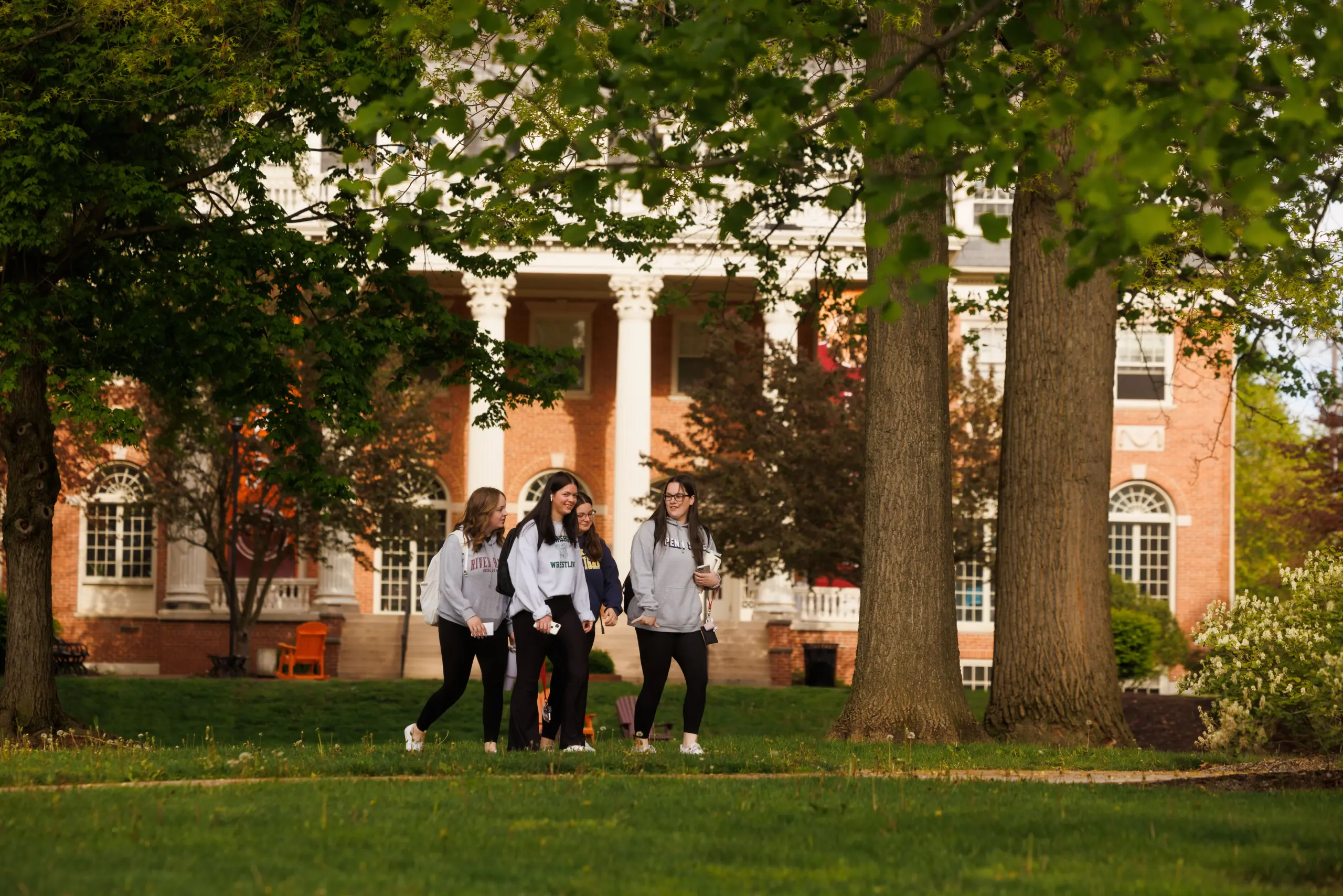 Four students walk on a green lawn surrounded by trees, with a red-brick building featuring white columns in the background. They appear to be chatting and are casually dressed, creating a relaxed, campus atmosphere.