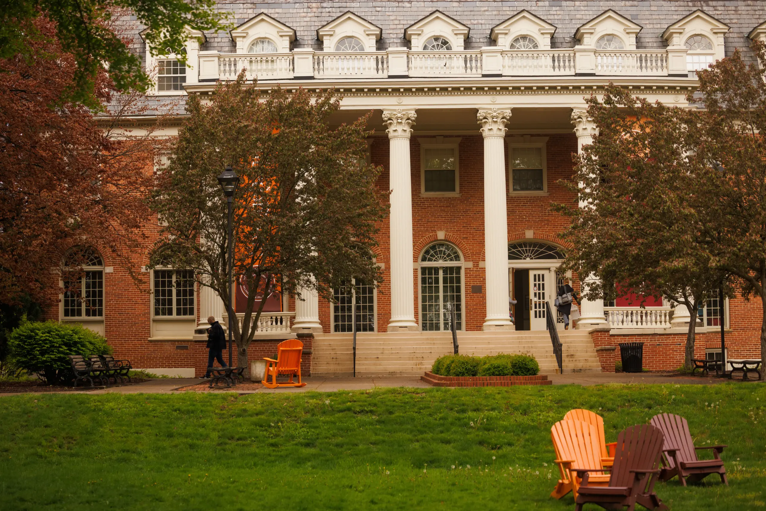 A brick building with white columns and arched windows stands amid lush greenery. Adirondack chairs in orange and brown sit on the lawn, with trees flanking the scene, adding a serene ambiance.