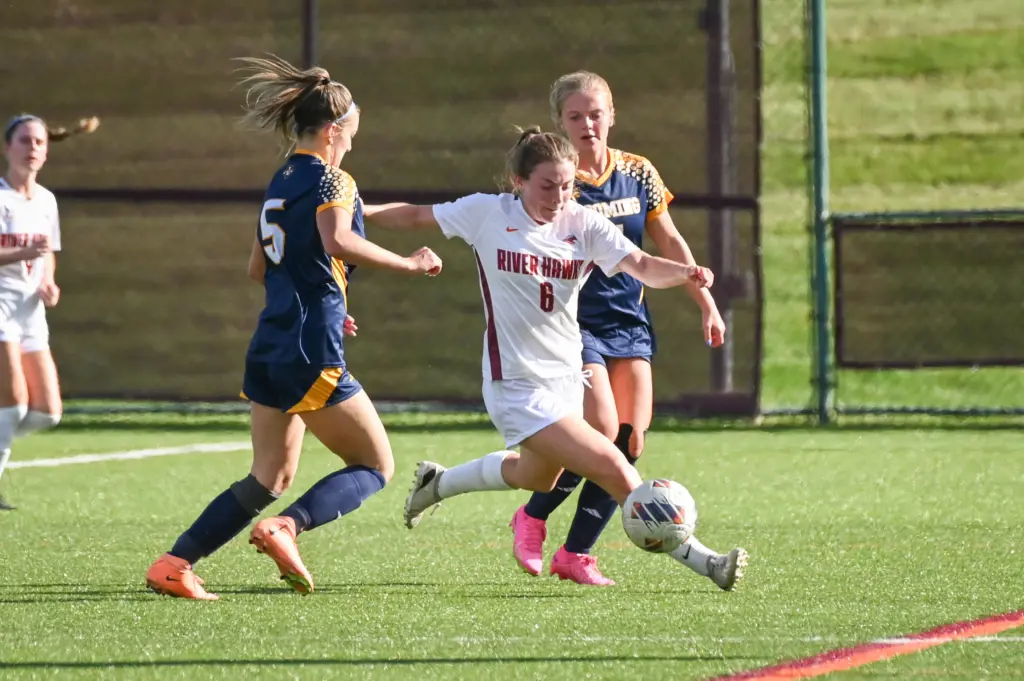 A soccer player in a white uniform attempts to kick the ball while two opponents in blue uniforms move in to block. The action takes place on a grassy field with a blurred background.