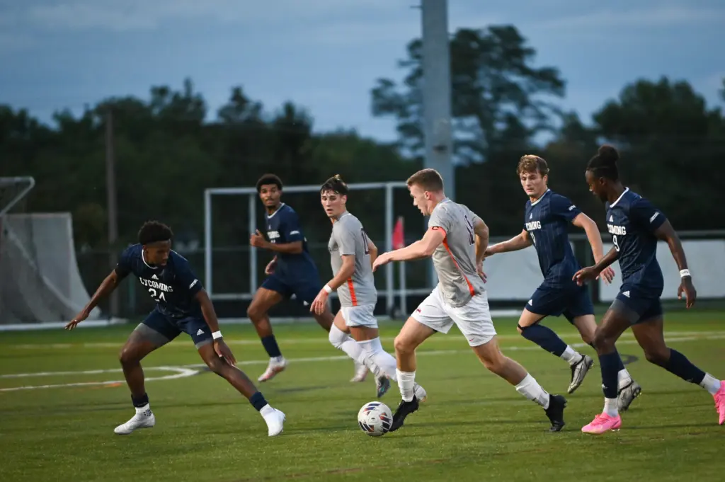 A soccer match in progress with a player in a white jersey dribbling the ball, closely pursued by several players in blue jerseys. The action takes place on a grassy field, with trees and a goalpost visible in the background.