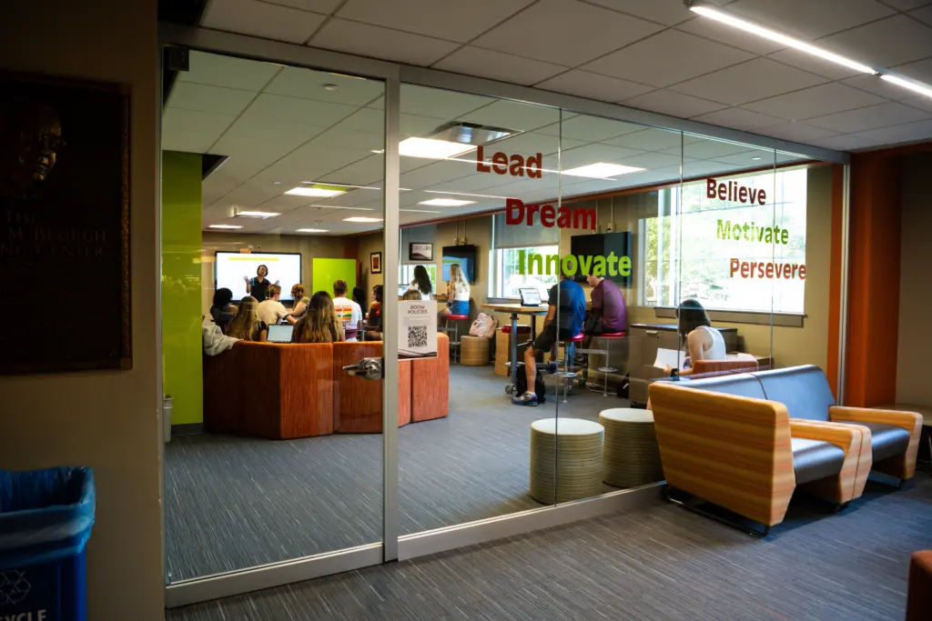 Students are gathered in a modern, glass-walled room with motivational words like "Lead," "Dream," and "Innovate" on the glass. The room has orange and striped seating, and a teacher is at the front near a whiteboard.