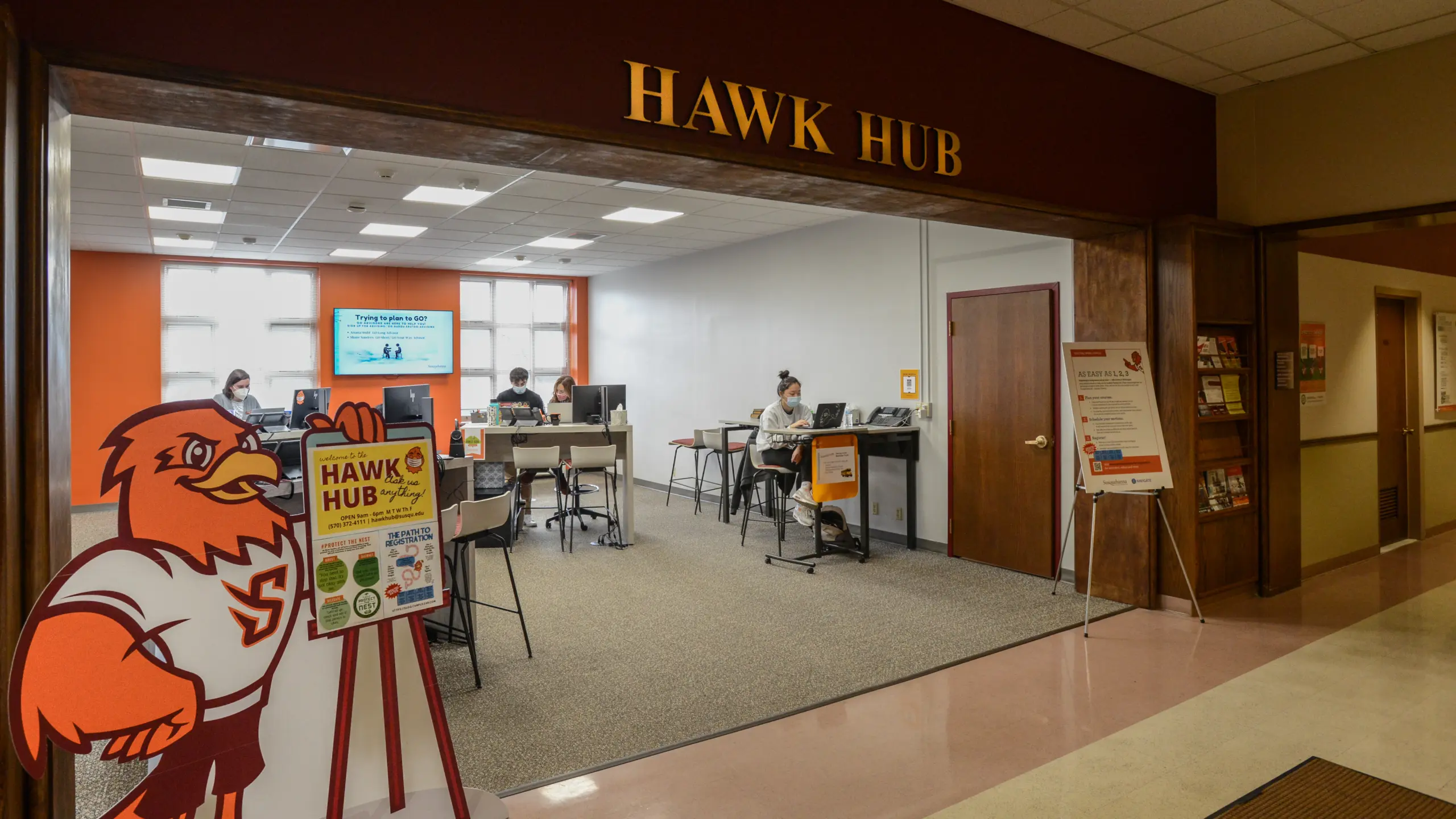 A room labeled "Hawk Hub" features a mascot cutout at the entrance. Inside, people sit at desks with computers in a modern, well-lit setting. Informational posters are displayed near the doorway, and a large screen is visible inside.