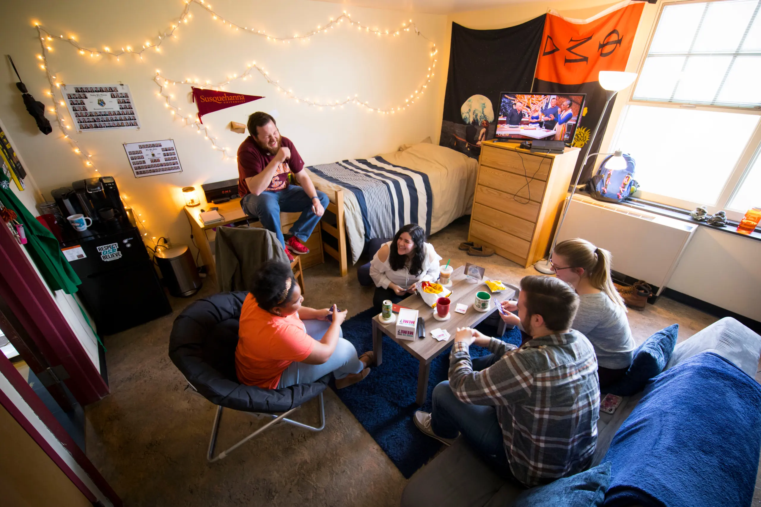 Five people are hanging out in a cozy college dorm room. The space is decorated with string lights and banners. A TV is on, and snacks are placed on the table as they chat and relax.
