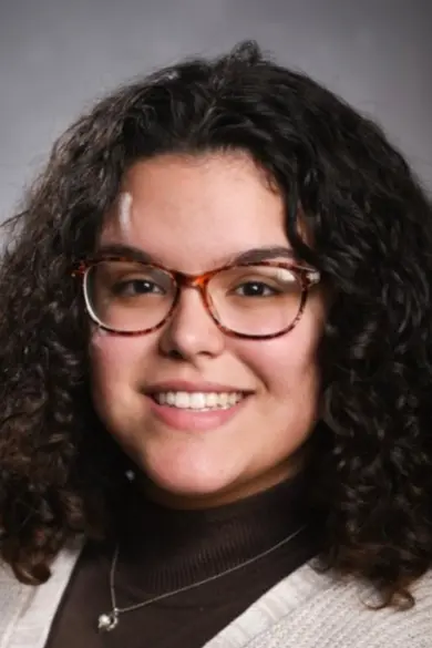Portrait of a person with curly dark hair, wearing glasses. They are smiling and wearing a brown turtleneck with a light sweater, against a neutral background.