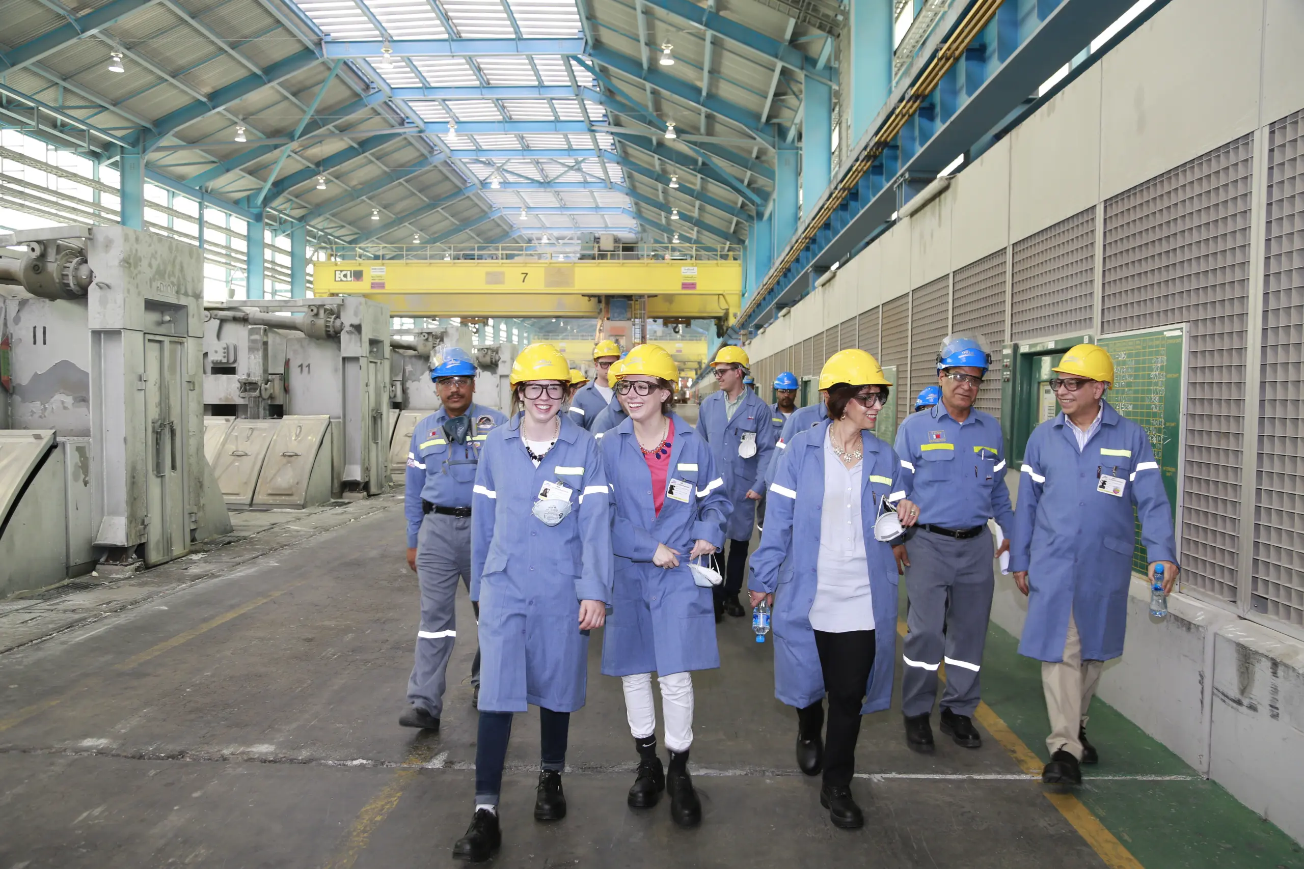 A group of people wearing blue industrial uniforms and yellow helmets walk through a large manufacturing facility. They appear to be on a tour or inspection, smiling and engaged in conversation as they move along the spacious, brightly lit space.