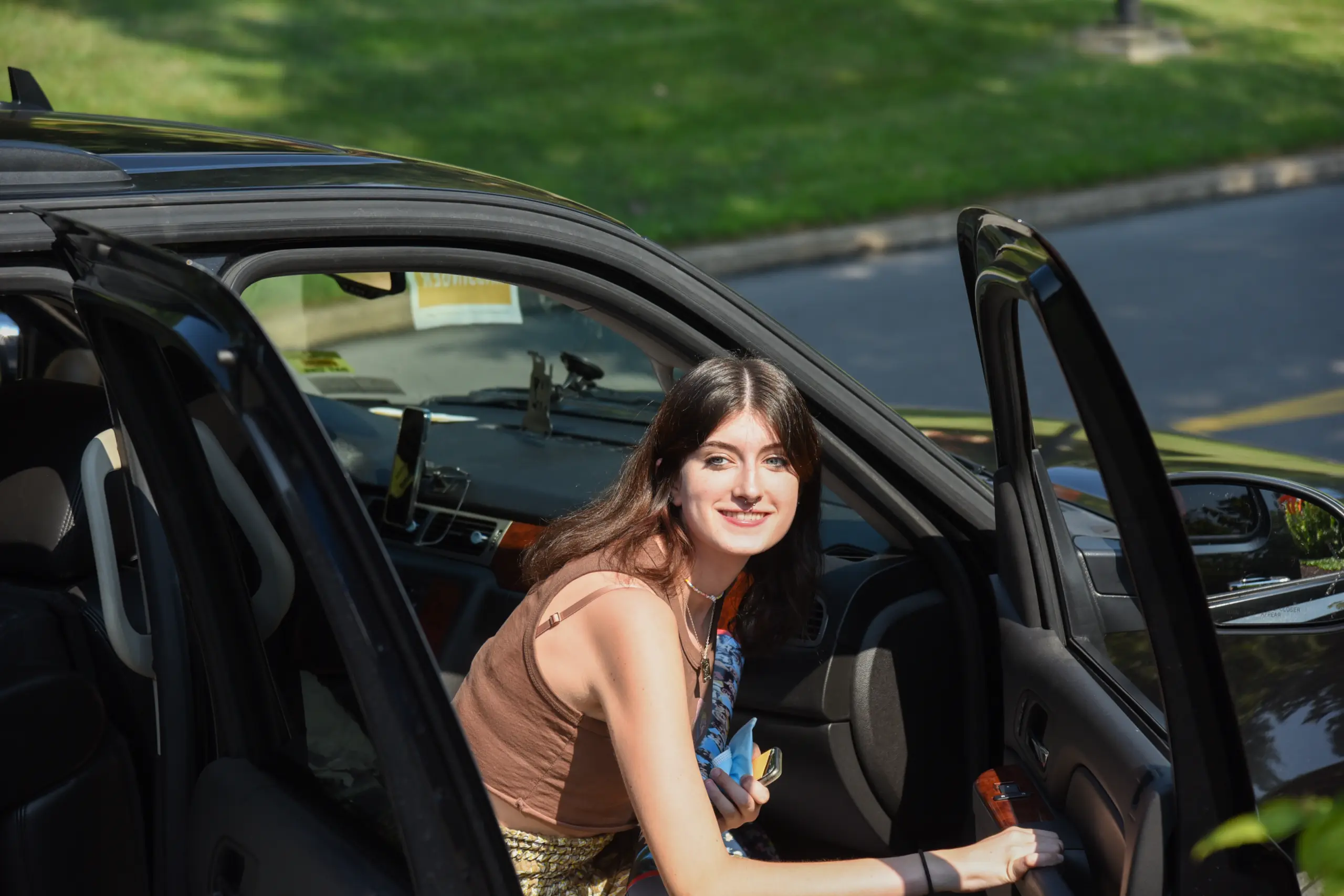 A woman with long dark hair sits in the passenger seat of a car with the door open, smiling at the camera. She is wearing a brown tank top and holding a small bouquet of flowers. Sunlit grass and pavement are visible in the background.