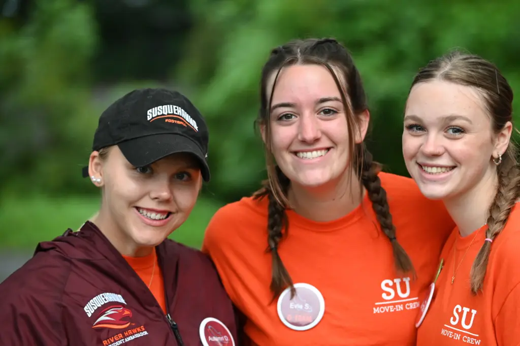 Three smiling individuals in orange shirts and a maroon jacket with "SU Move-In Crew" logos. One wears a black cap. They are outdoors, and trees are blurred in the background.