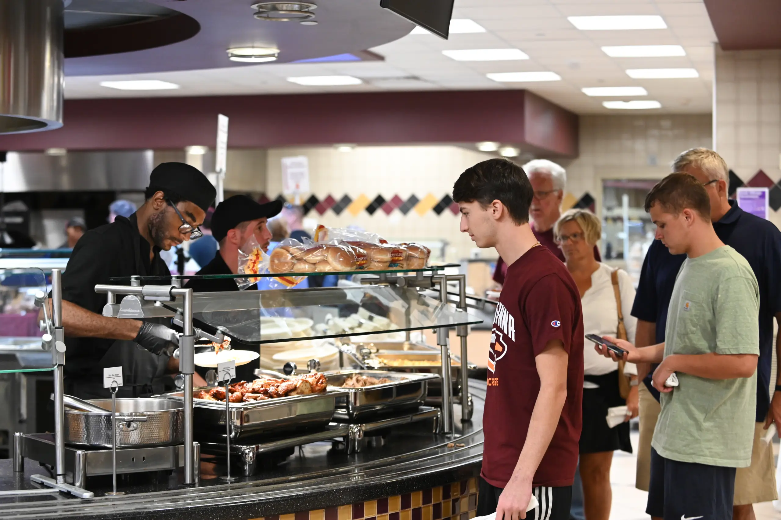 People line up at a cafeteria-style food counter where a staff member, wearing a cap and gloves, serves food. Two young men in casual clothing wait at the front of the line, and more customers stand behind them.