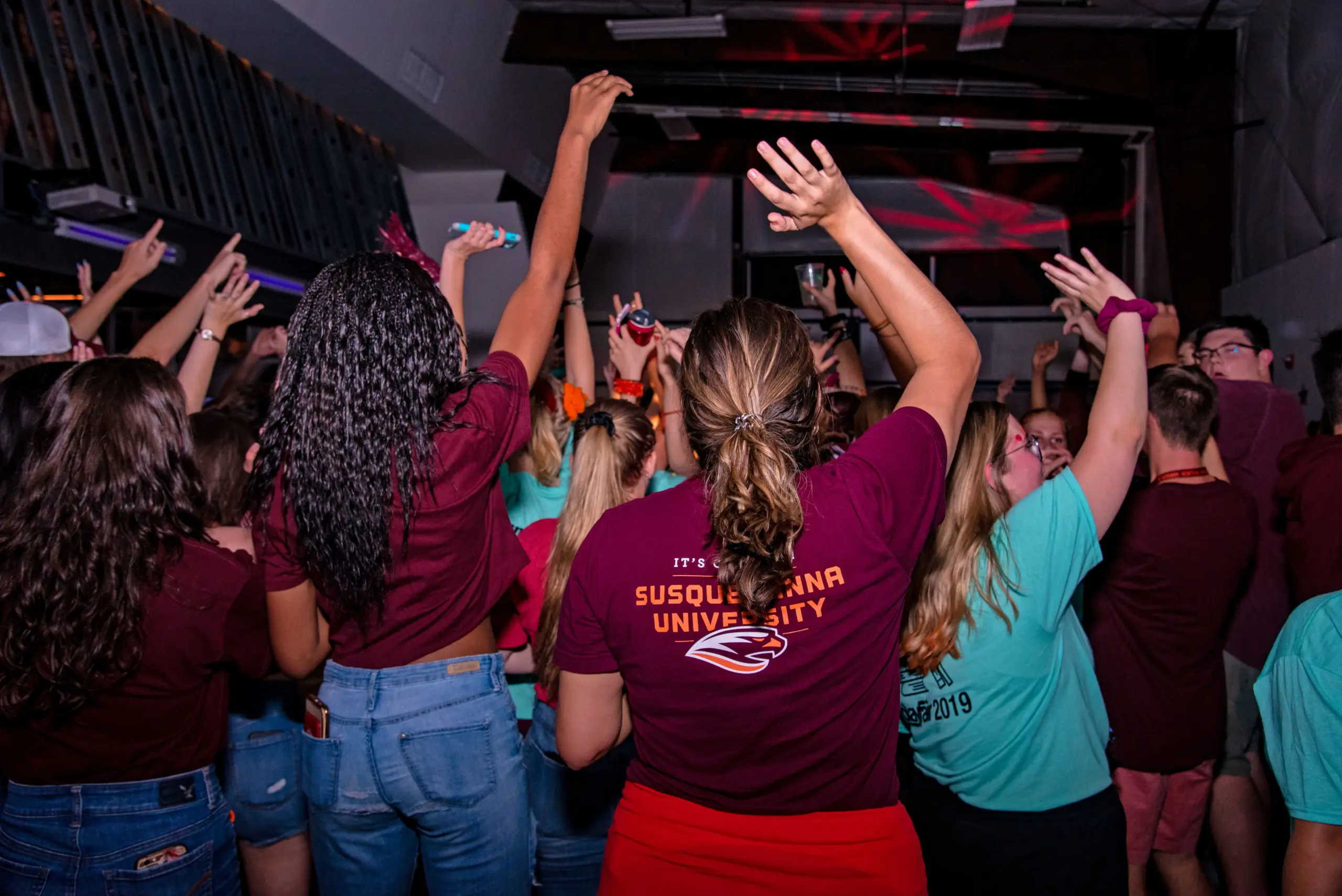 A lively crowd of people in casual wear dance and raise their hands at an indoor event. Several individuals are wearing maroon and turquoise shirts, with one shirt displaying "Susquehanna University." Colorful lights illuminate the scene.
