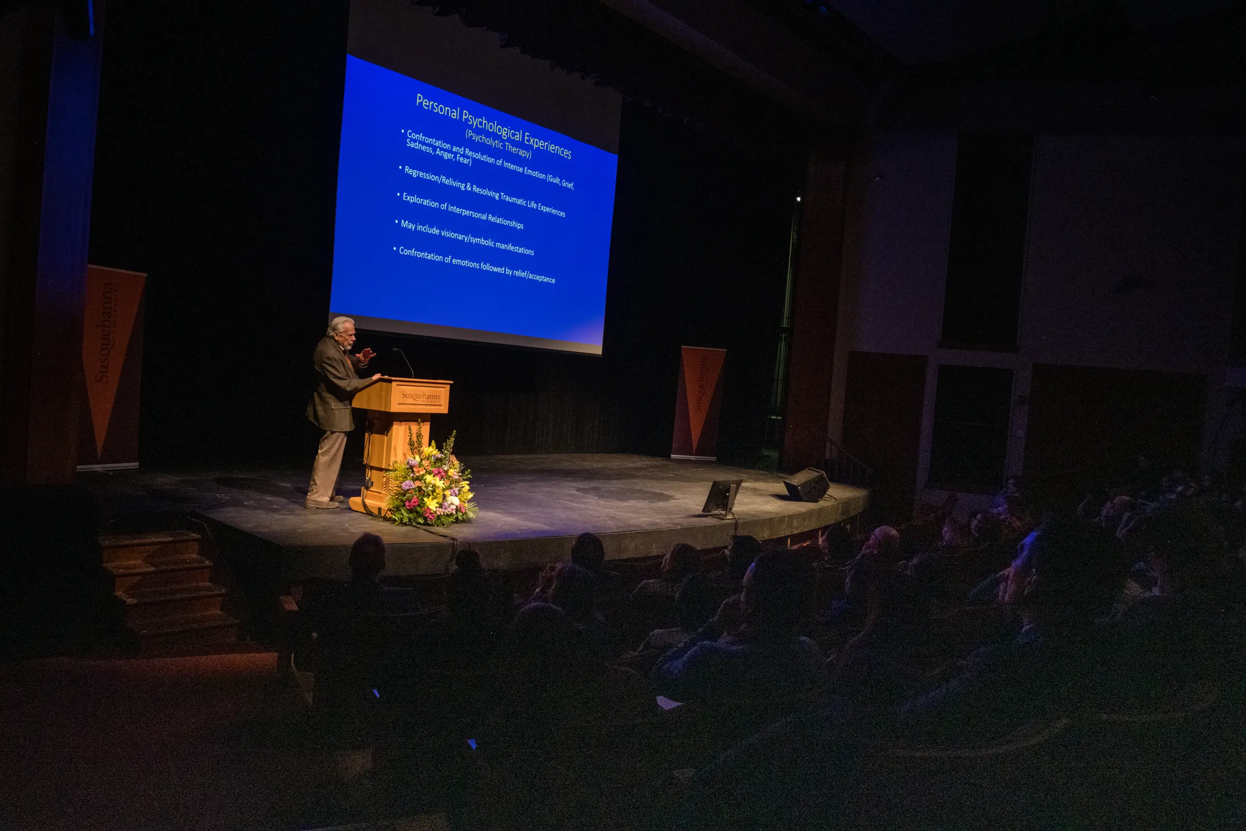 A person stands on a stage giving a presentation in a dimly lit auditorium. The slide displayed is titled "Personal Psychological Experience." The audience is seated in rows, listening attentively. A podium and floral arrangement are visible on the stage.