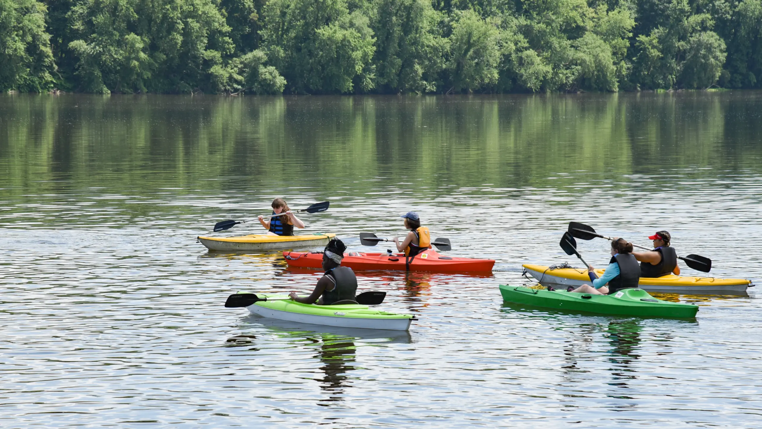 Five people are kayaking on a calm lake surrounded by lush green trees. They are paddling in colorful kayaks, including yellow, red, and green, under a clear sky. The serene water reflects the trees and kayakers.