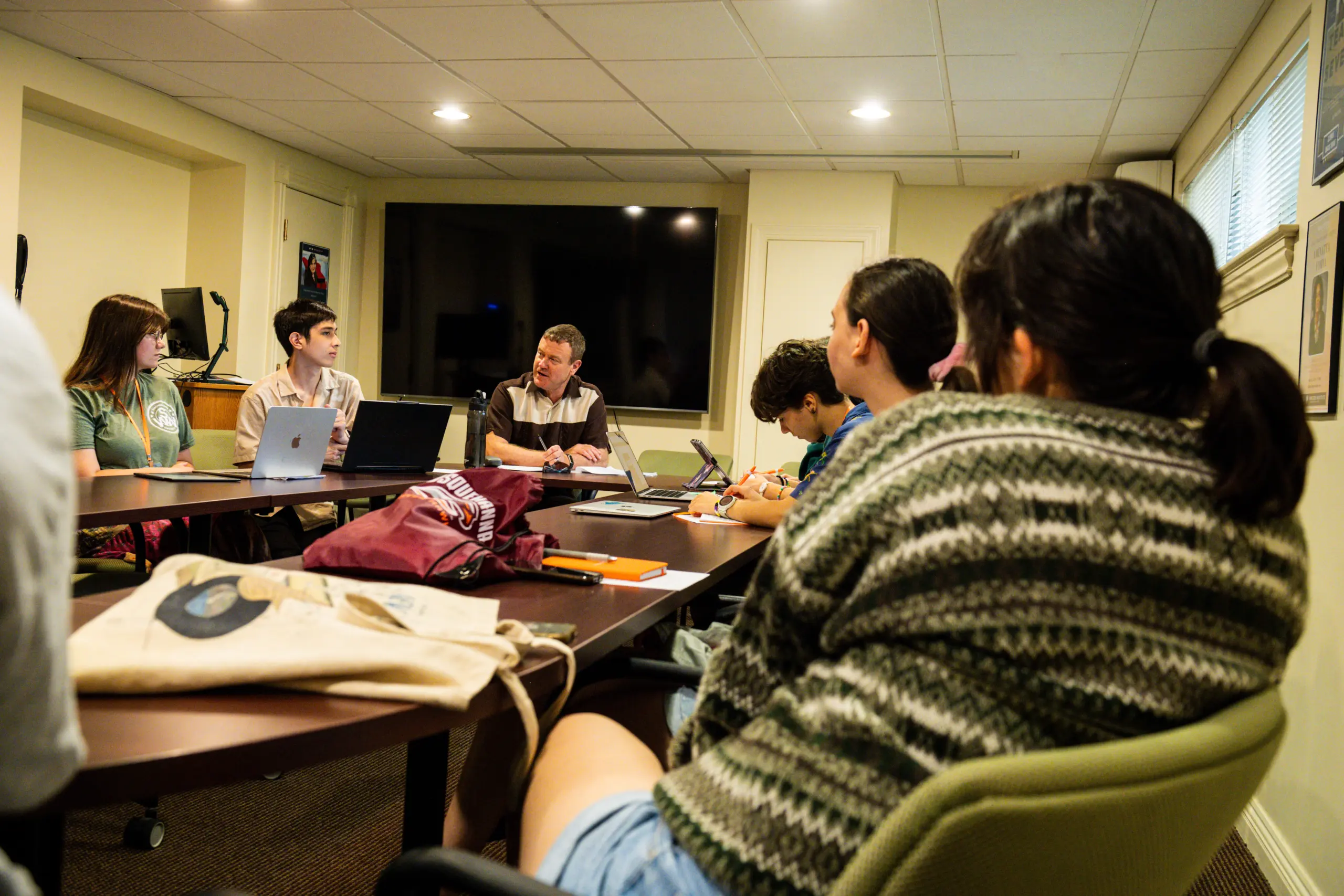 A group of people sits around a conference table in a meeting room, engrossed in a Writers Workshop. Laptops and notebooks are scattered on the table. A large television is mounted on the wall, enhancing their collaborative efforts under the room’s bright overhead lighting.