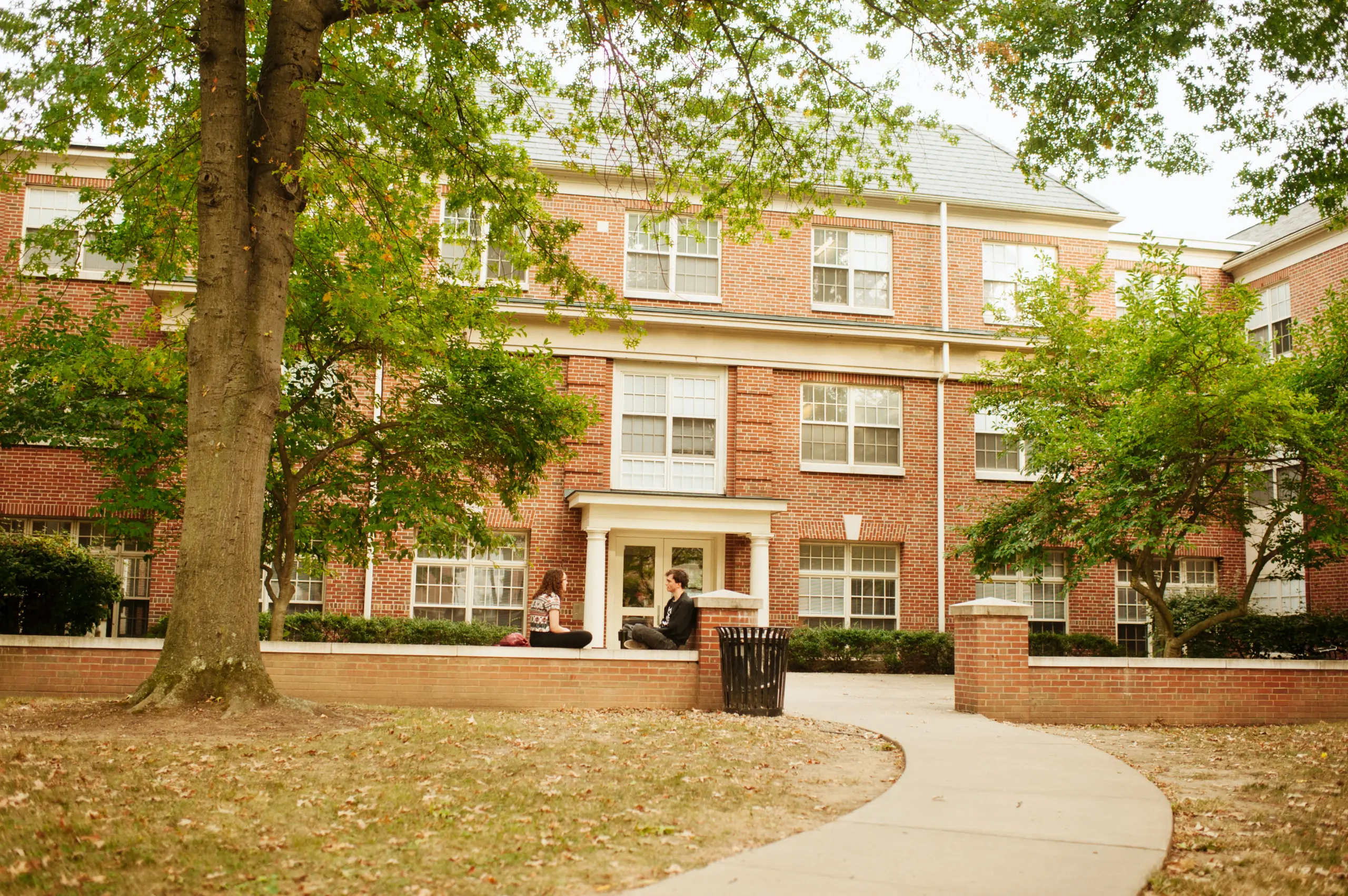 Three people sit on a low brick wall in front of a large brick building with white-trimmed windows and a central entrance. The scene is framed by trees and a curved pathway leading to the building.