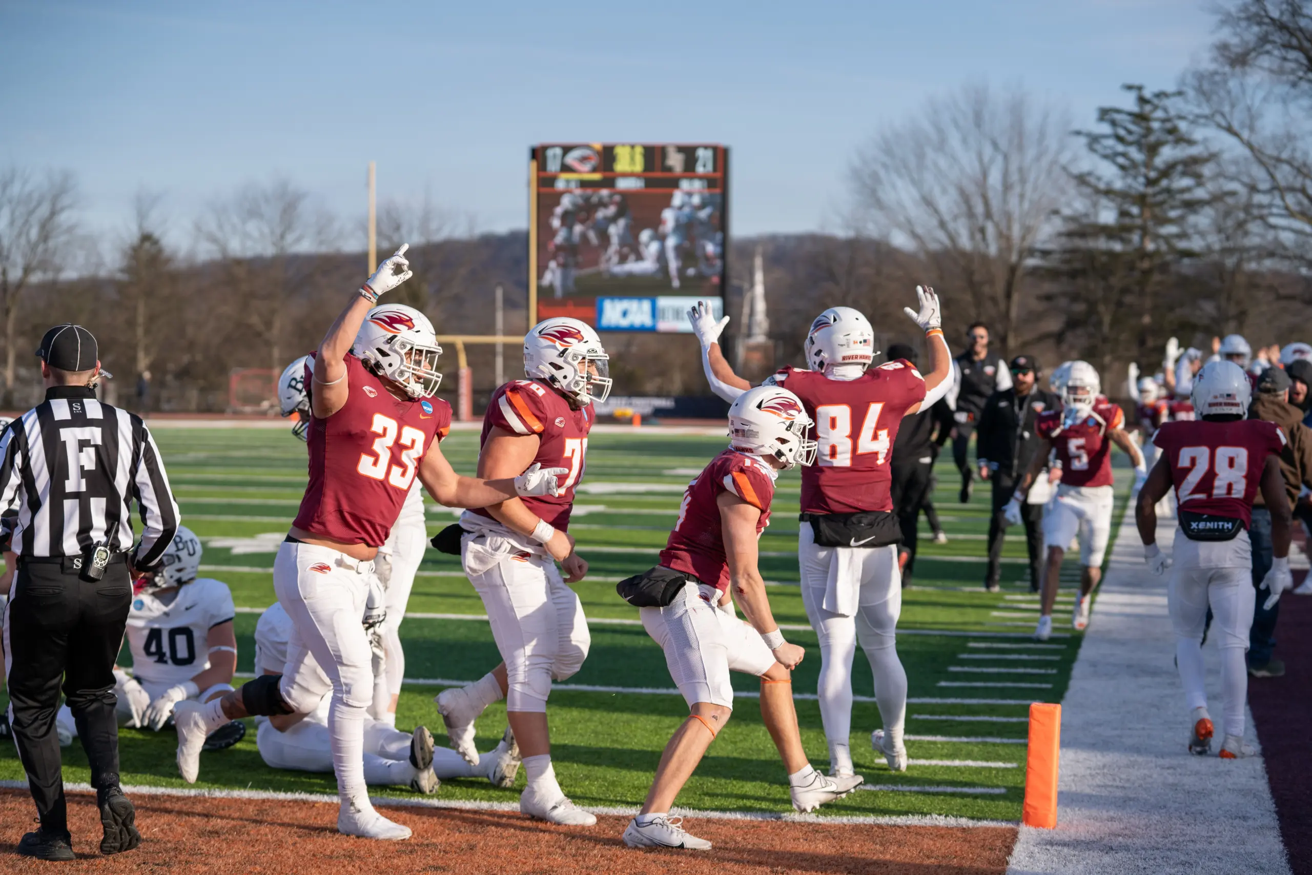 Football players in red and white uniforms celebrate near the sideline on a sunny day. A referee stands nearby, and other players are visible in the background. A scoreboard is seen in the distance.