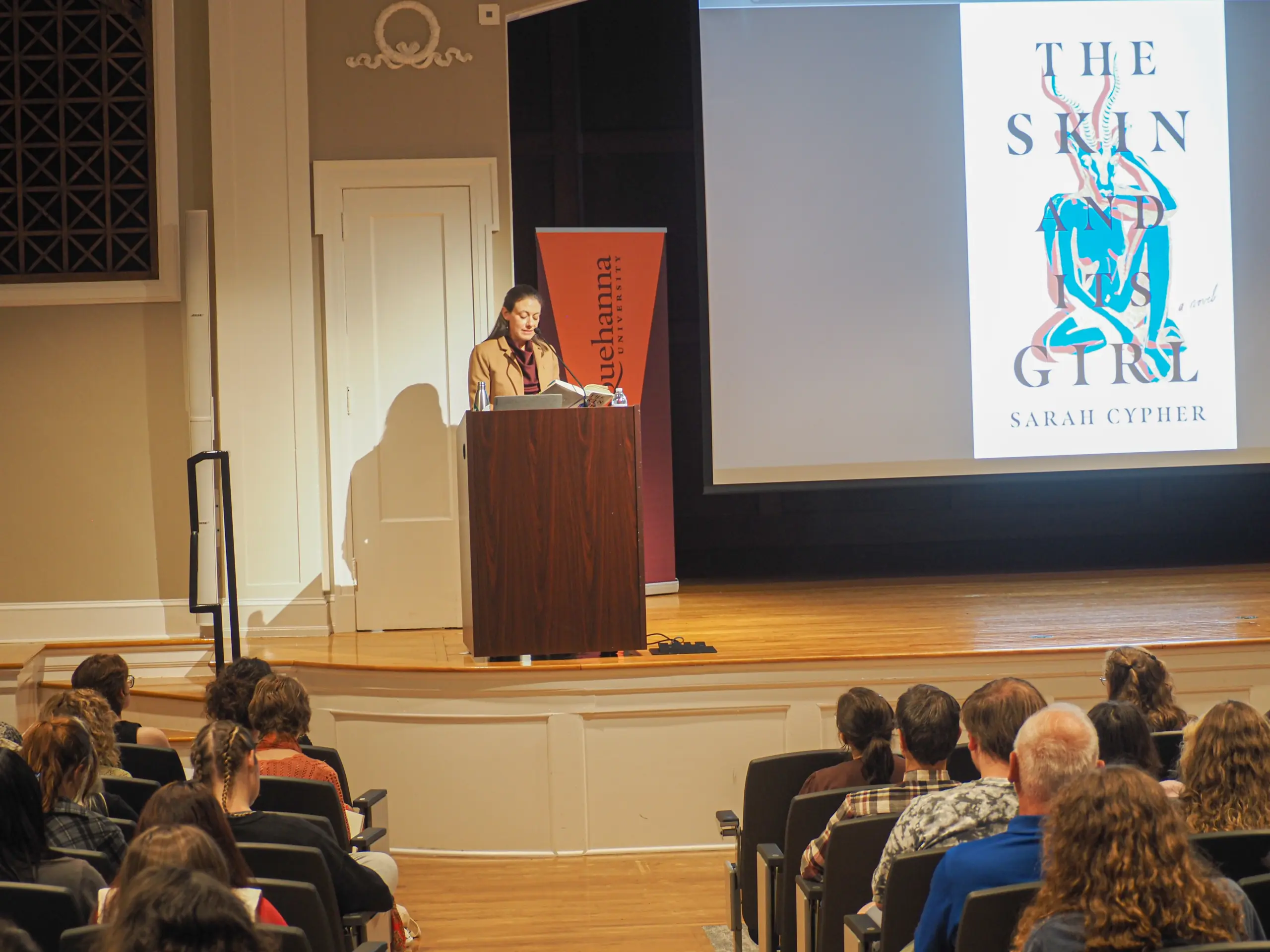 A person stands at a podium giving a lecture or presentation to an audience. On a screen, the book cover "The Skin and Its Girl" by Sarah Cypher is displayed. The room is well-lit and filled with seated attendees.