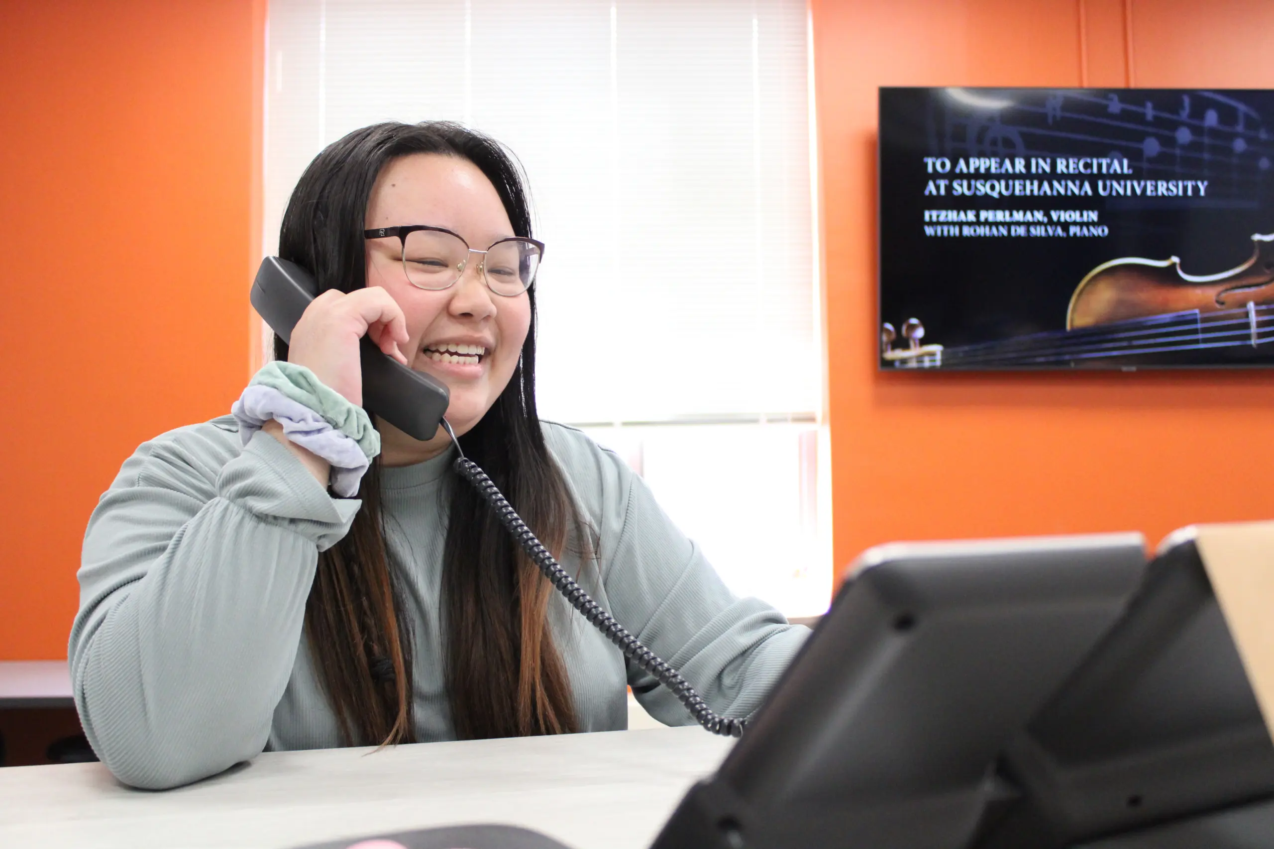 A smiling person with long dark hair and glasses chats on a landline phone in an office with orange walls, likely discussing student employment opportunities. A poster on the wall announces a recital at Susquehanna University.