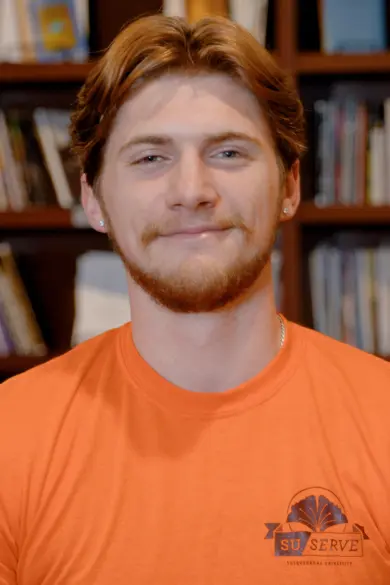 A person with short, light brown hair and a beard smiles slightly while wearing an orange shirt that says "SU Serve." They are standing in front of a blurred bookshelf.
