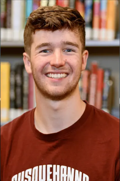 A smiling individual with short brown hair wearing a maroon shirt stands in front of a blurred background of books on shelves.