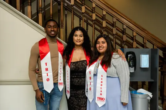 Three people are standing together, wearing red and white stoles labeled "TRIO Scholar." They are posing in front of a wooden staircase, smiling and dressed in formal attire.