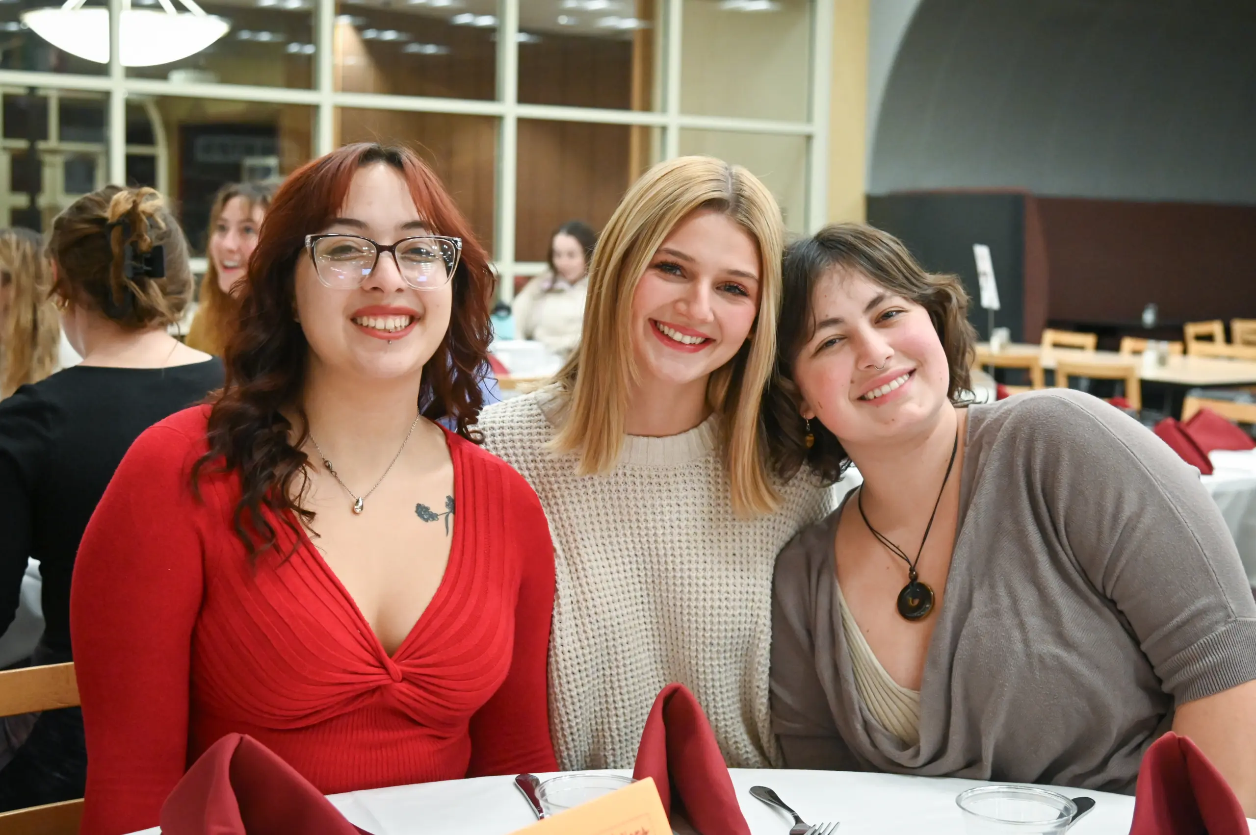 Three smiling people sit at a table with white tablecloths and red napkins. The person on the left wears glasses and a red top, the middle person has a cream sweater, and the person on the right wears a gray top. The background shows more people and dining hall decor.
