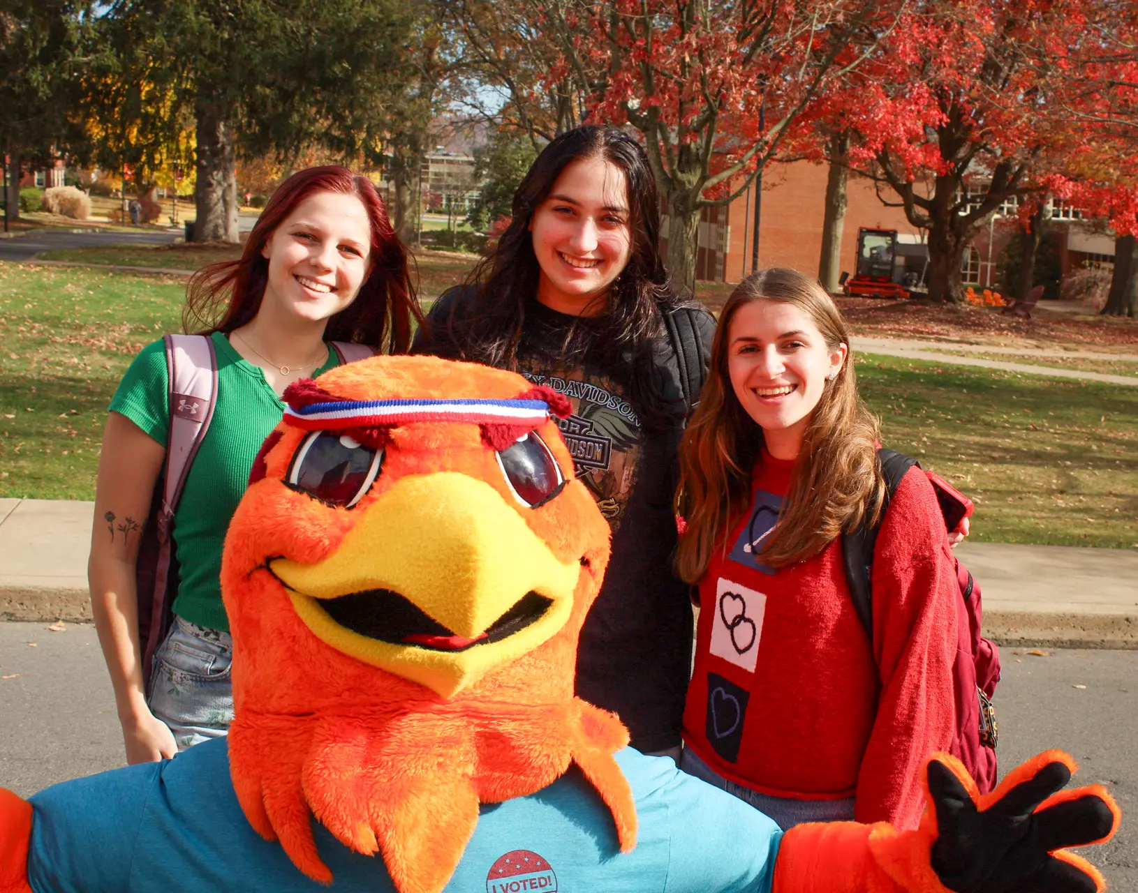 Three smiling people stand outdoors with a colorful bird mascot in front of them. They are in a park-like setting with autumn trees in the background. The mascot wears a blue shirt with a sticker that says "I Voted.