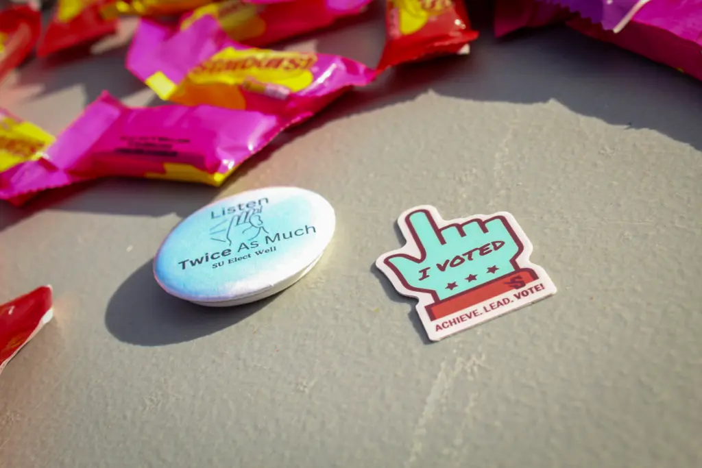 A button reading "Listen Twice As Much" and a sticker saying "I Voted" with "Achieve, Lead, Vote" are placed on a table. Colorful candy wrappers are scattered in the background.
