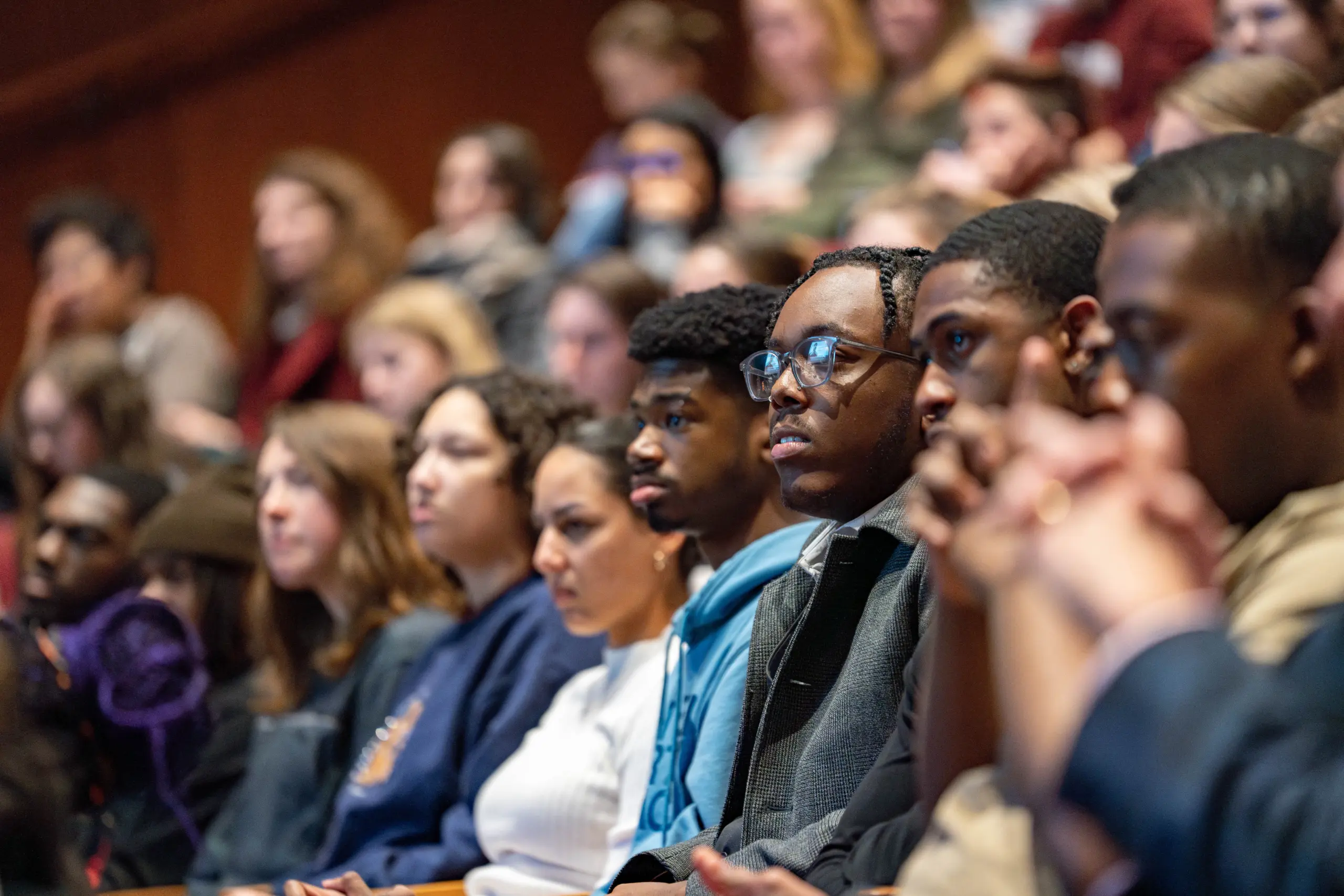 A diverse group of people seated closely in an auditorium, attentively watching a presentation. The audience includes individuals of different ages and backgrounds, all focused on the speaker or event happening off-camera.