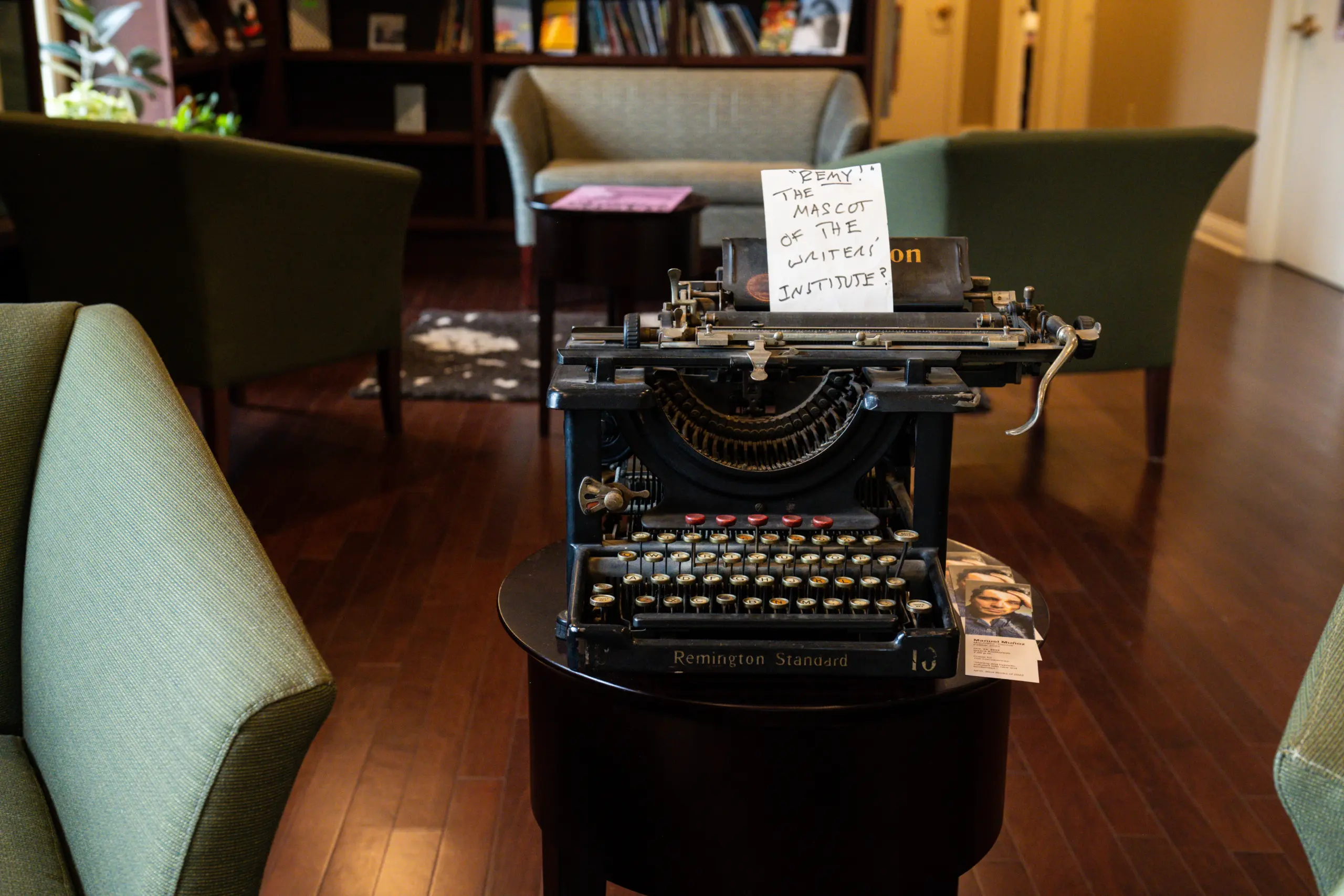 A vintage Remington Standard typewriter sits proudly on a small table, fusing charm with nostalgia in a cozy room. With wooden floors and green armchairs, the sign reads, "Larry: The Mascot of the Writers' Institute." Books line the shelves, completing the inviting scene.