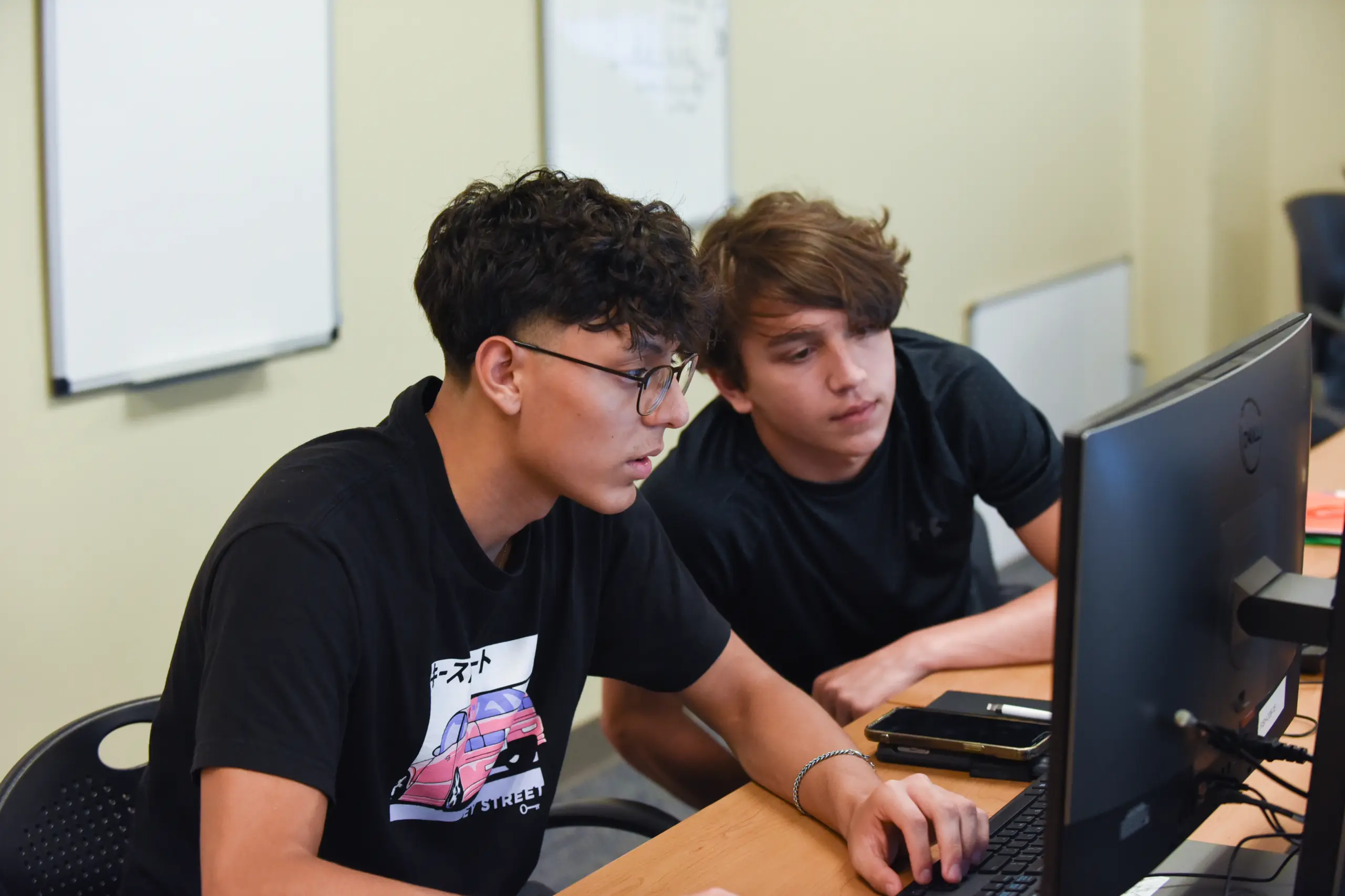 Two young men, both in black shirts, focus intently on a computer screen. One is typing while the other observes. They are seated at a desk with whiteboards in the background.