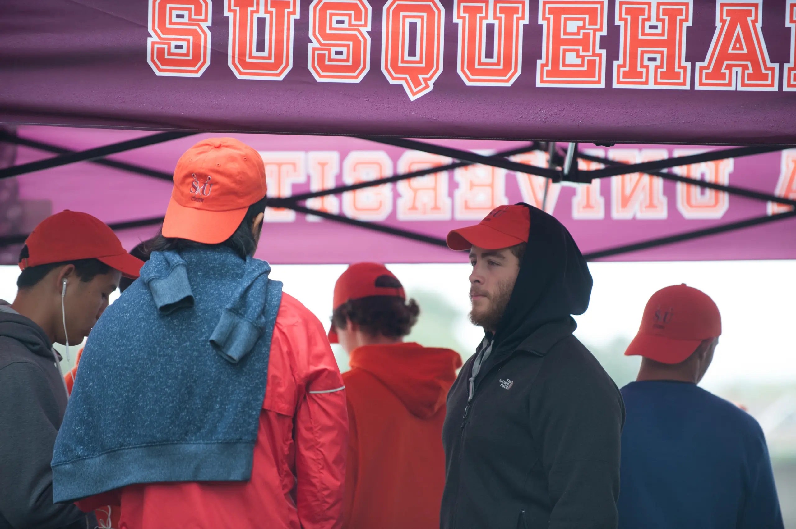 Several people wearing red hats and jackets stand under a purple canopy with "SUSQUEHANNA" partially visible in orange letters. One person wears a hoodie and another has a towel draped over their shoulders.