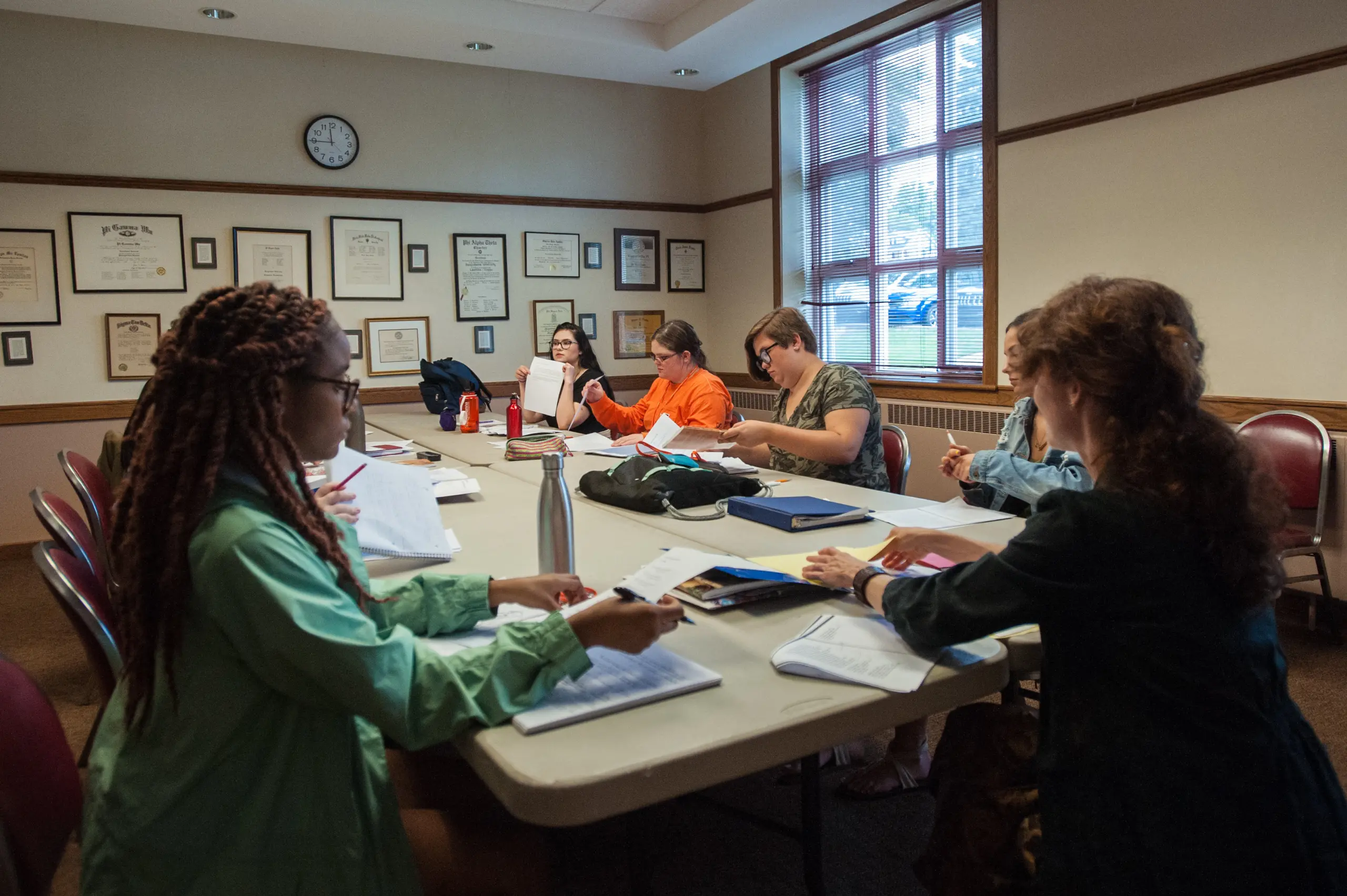 A group of people of various genders sit around a table in a conference room, engaged in discussion and taking notes. The table is covered with papers, notebooks, and personal items. Certificates hang on the wall, and a window is in the background.