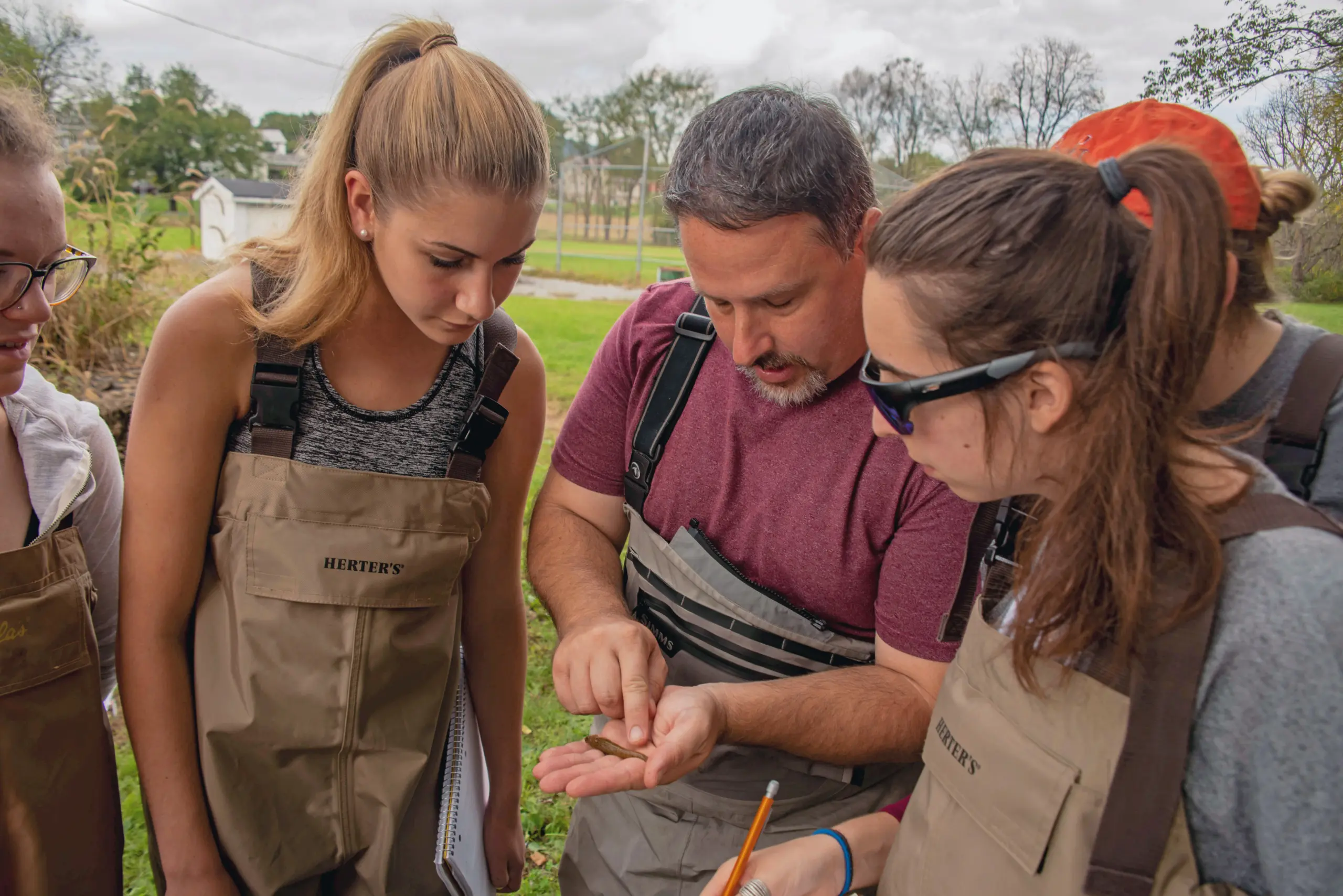 A group of people wearing waders examine an object closely in a man's hand. The man gestures while others observe intently. Trees and grass are visible in the background, suggesting an outdoor setting.