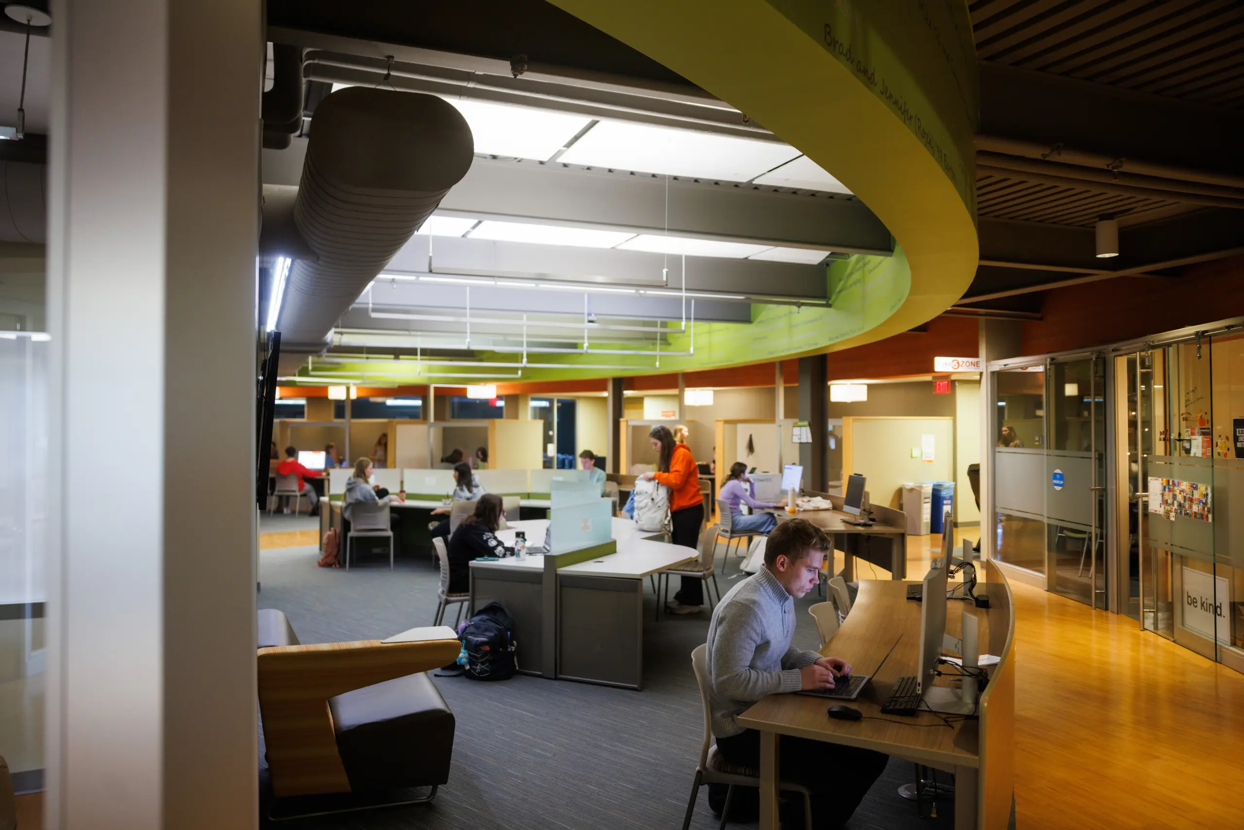 A spacious, modern office with people working at desks and cubicles. The room features bright lighting, curved architectural elements, and glass walls. Several individuals are using laptops, and the atmosphere appears focused and professional.