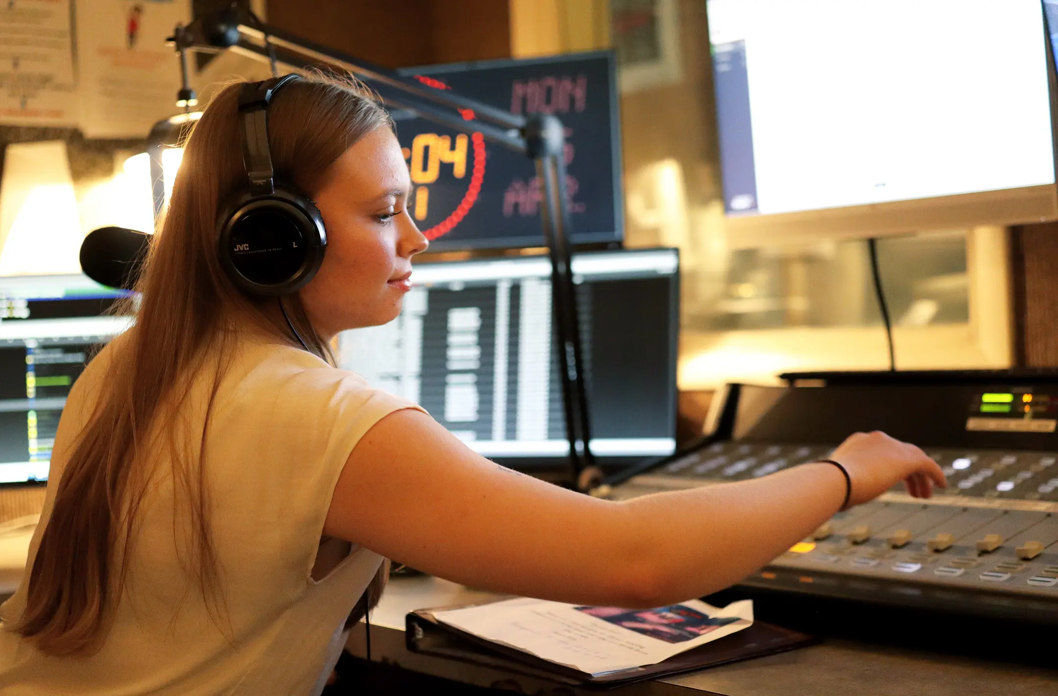 A woman with long hair is wearing headphones and operating a sound mixing board in a radio studio. As she deftly adjusts the controls, surrounded by monitors and a partially visible clipboard, her expertise enhances the station's student publications.