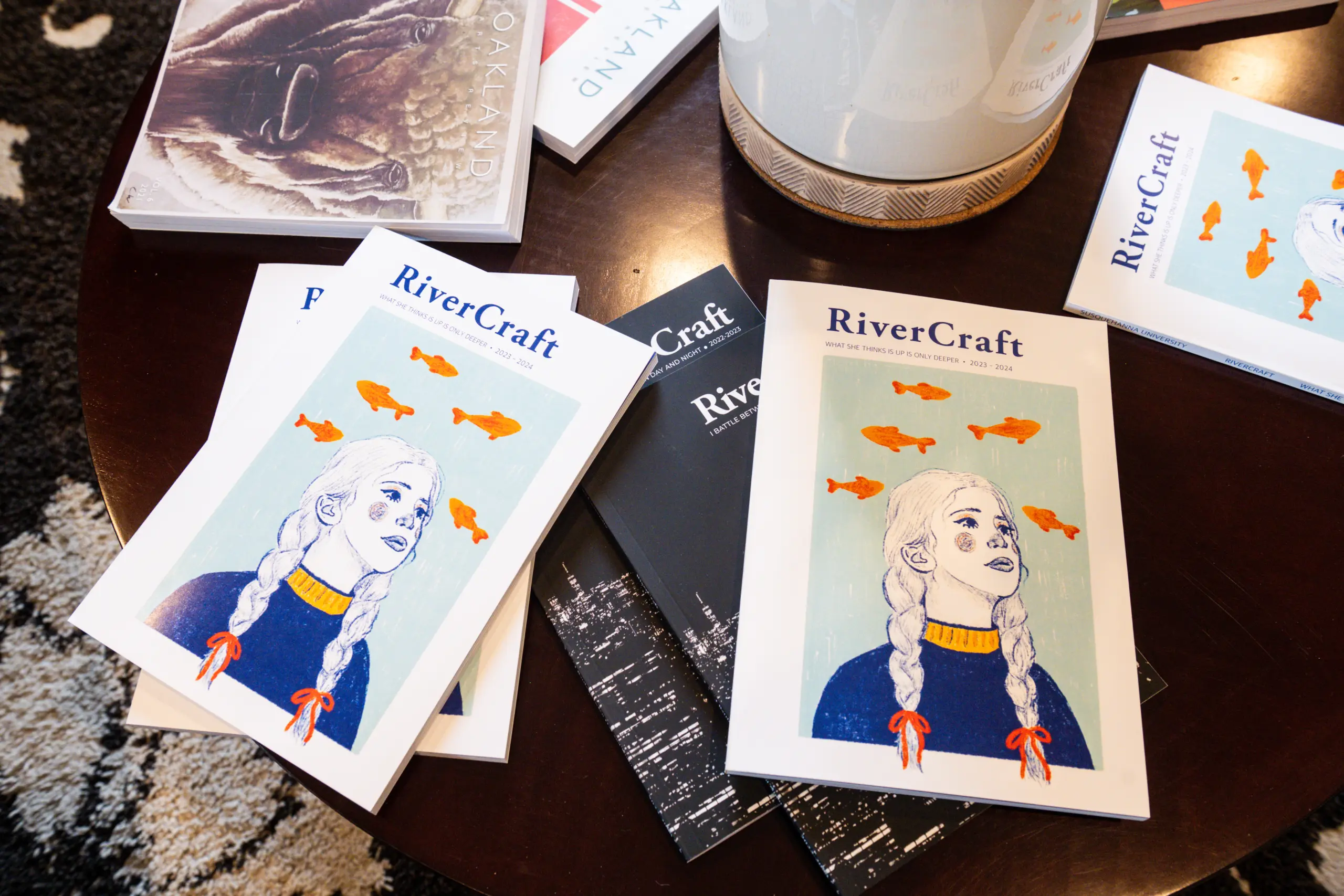 A table displays several copies of "RiverCraft" magazine, a standout in student publications. The cover features a person with braided hair surrounded by orange fish, while other books and decorative items create a vibrant backdrop.