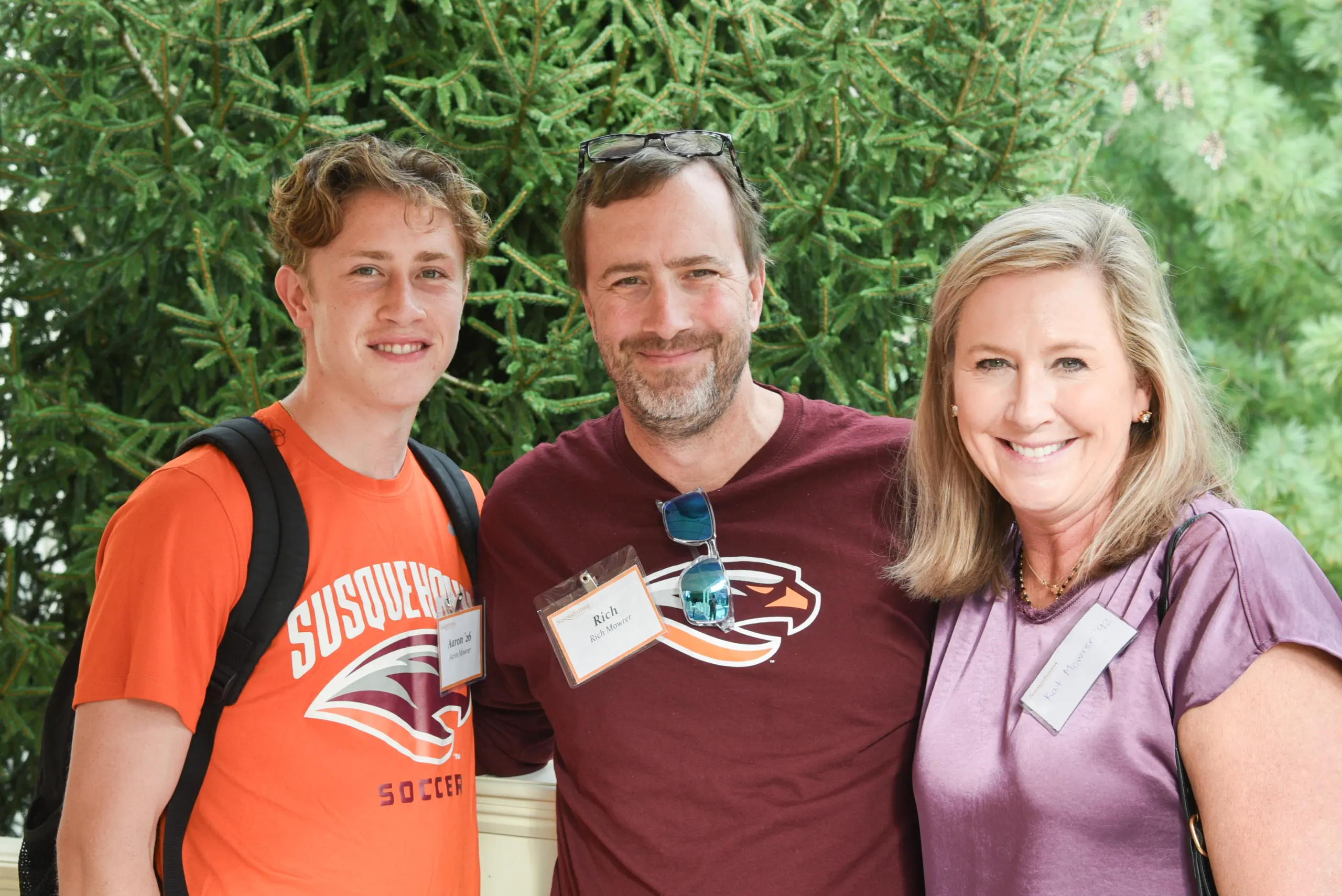 Three people attend a gathering, smiling in front of lush greenery. The person on the left wears an orange shirt, the person in the middle sports a dark shirt and glasses, and the one on the right dons a purple blouse. All proudly display their name tags.