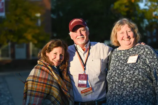 Three people are smiling outdoors in a sunny setting. The person in the middle, symbolizing "Ways to Give," wears a red cap and lanyard. On the left, someone is wrapped in a plaid shawl, while on the right, there's another dressed in a blue-patterned top. Lush trees stand tall in the background.