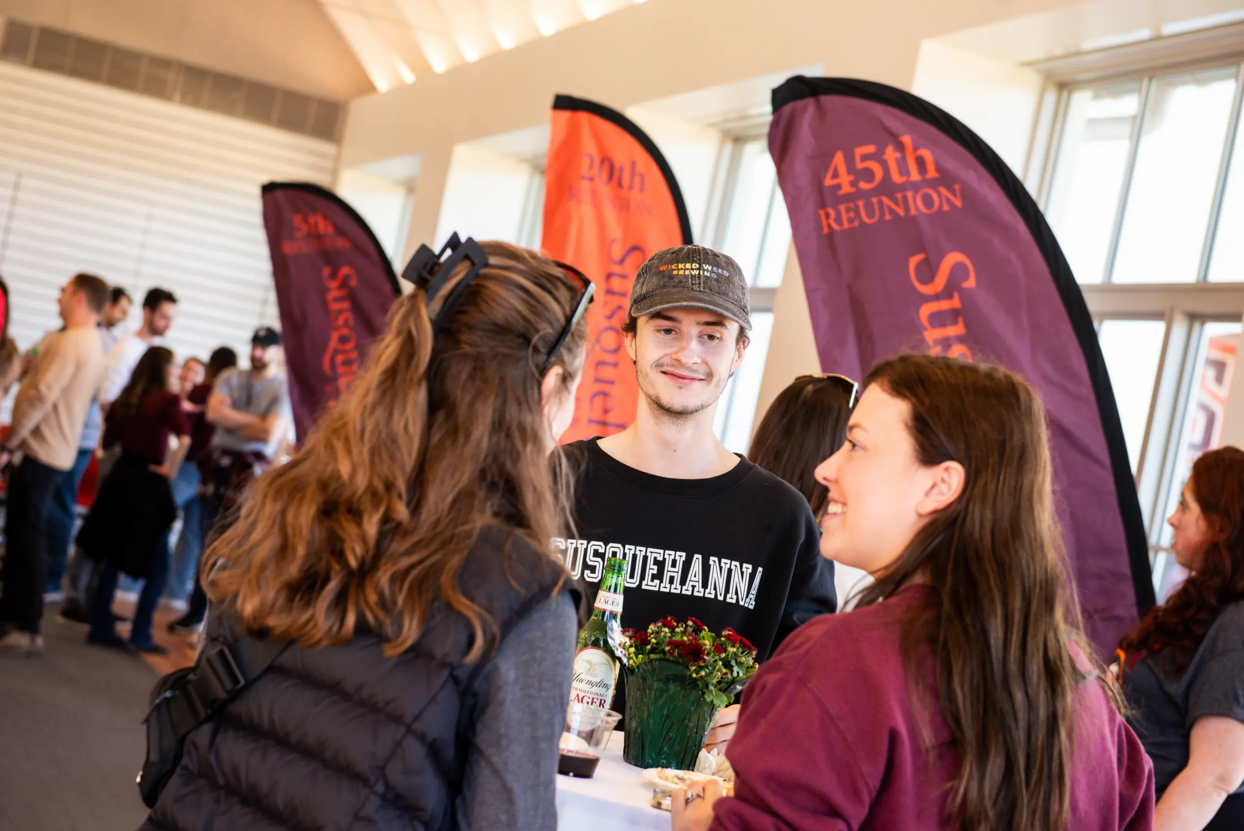 A group of people are gathered in a room for a joyful homecoming reunion, talking and smiling. They stand near colorful banners, one reading "45th Reunion." The setting is indoors with bright natural lighting streaming through large windows.