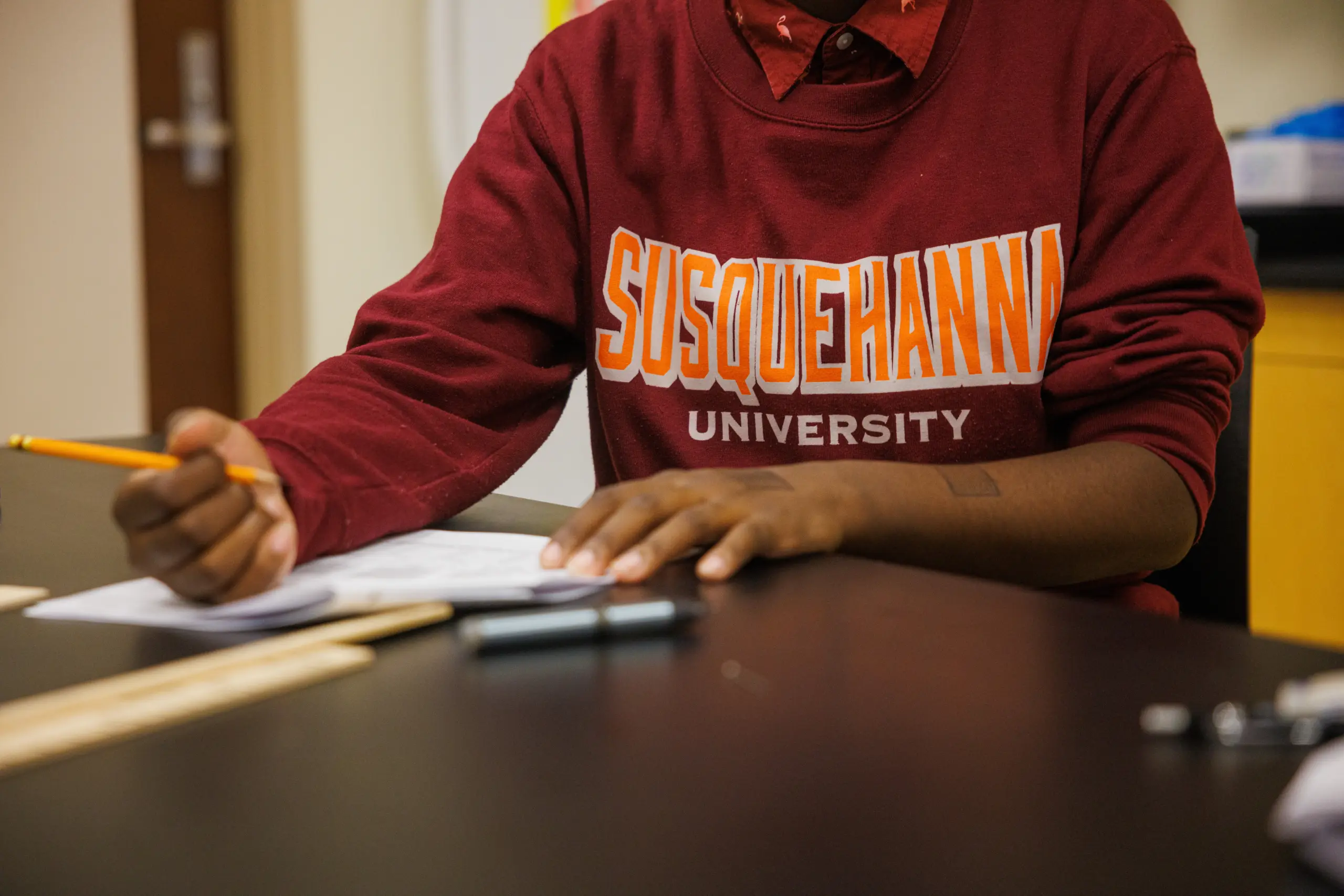 Person wearing a maroon "Susquehanna University" sweatshirt, writing with a pencil at a desk. The table has papers and a ruler. The background shows an office environment.