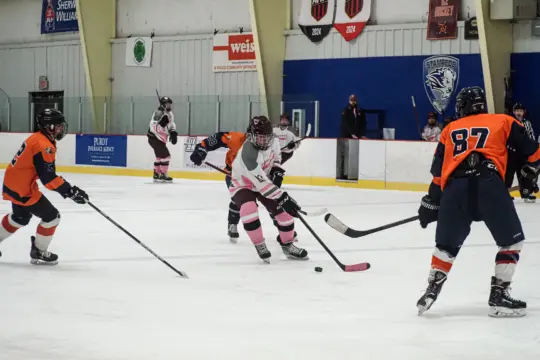 Hockey players in orange and black jerseys compete against those in white and pink on an ice rink. Engaging in exhilarating athletics, one player in white skillfully skates with the puck while others defend. Spectators and colorful banners enhance the vibrant recreation setting.