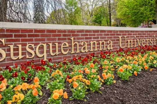 A brick wall with "Susquehanna University" in large letters. In front, there is a flower bed with orange and red flowers. Trees with spring foliage are visible in the background.