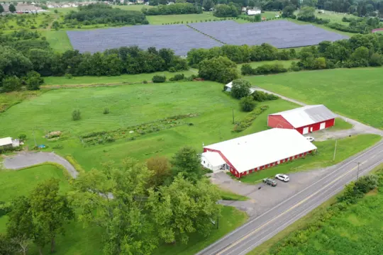 Aerial view of a rural landscape featuring a large red barn with a white roof, surrounded by lush green fields. A paved road runs alongside the barn. In the background, a field with rows of solar panels is visible. Trees and greenery are scattered throughout.