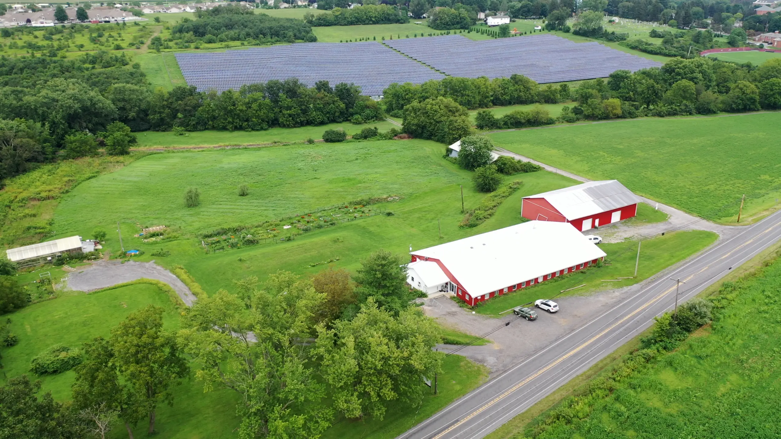 Aerial view of a rural landscape featuring a large red barn with a white roof, surrounded by lush green fields. A paved road runs alongside the barn. In the background, a field with rows of solar panels is visible. Trees and greenery are scattered throughout.