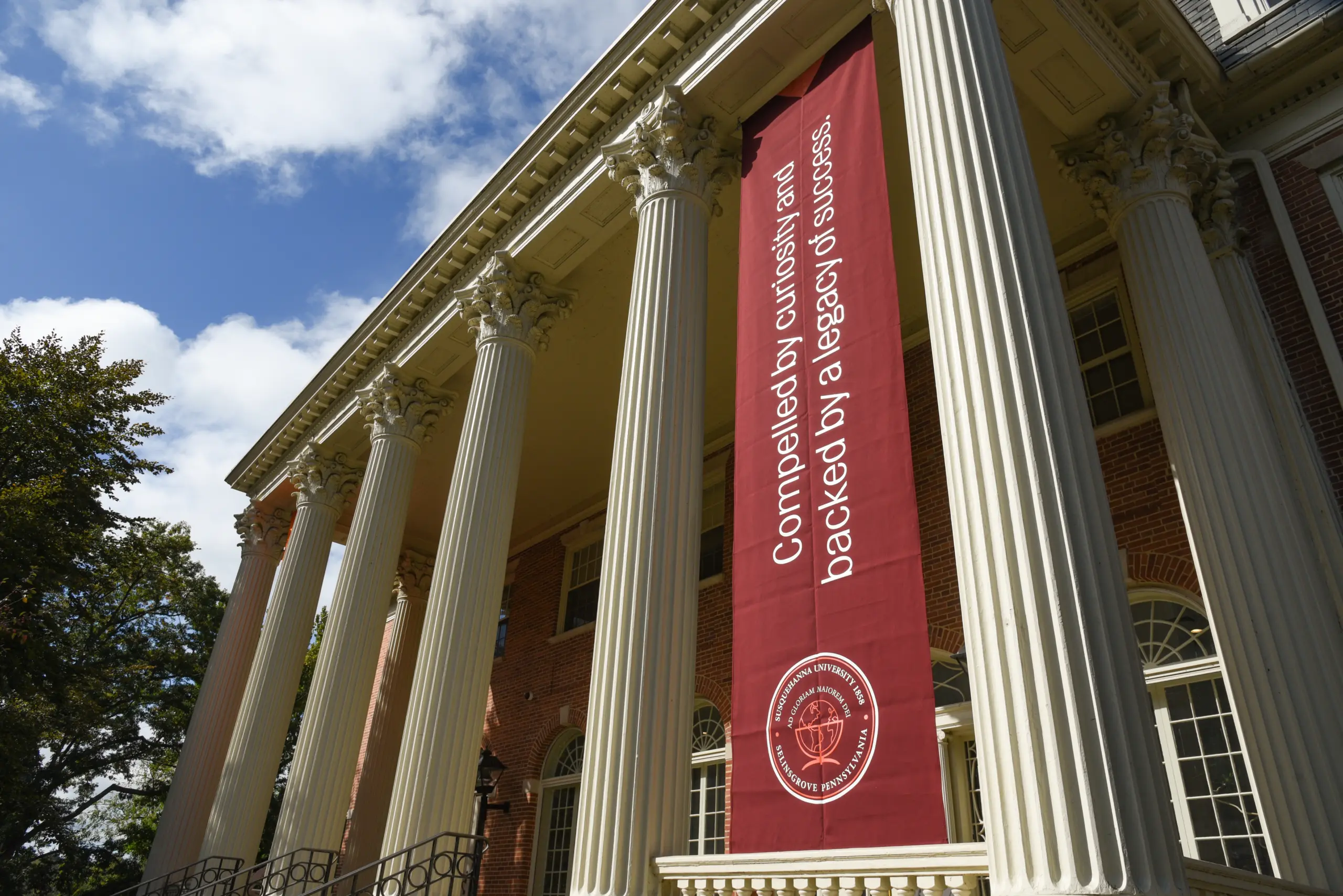A historic building with large columns and a red banner hanging between them. The banner reads, "Compelled by curiosity and backed by a legacy of successes," with an emblem at the bottom. The sky is partly cloudy.